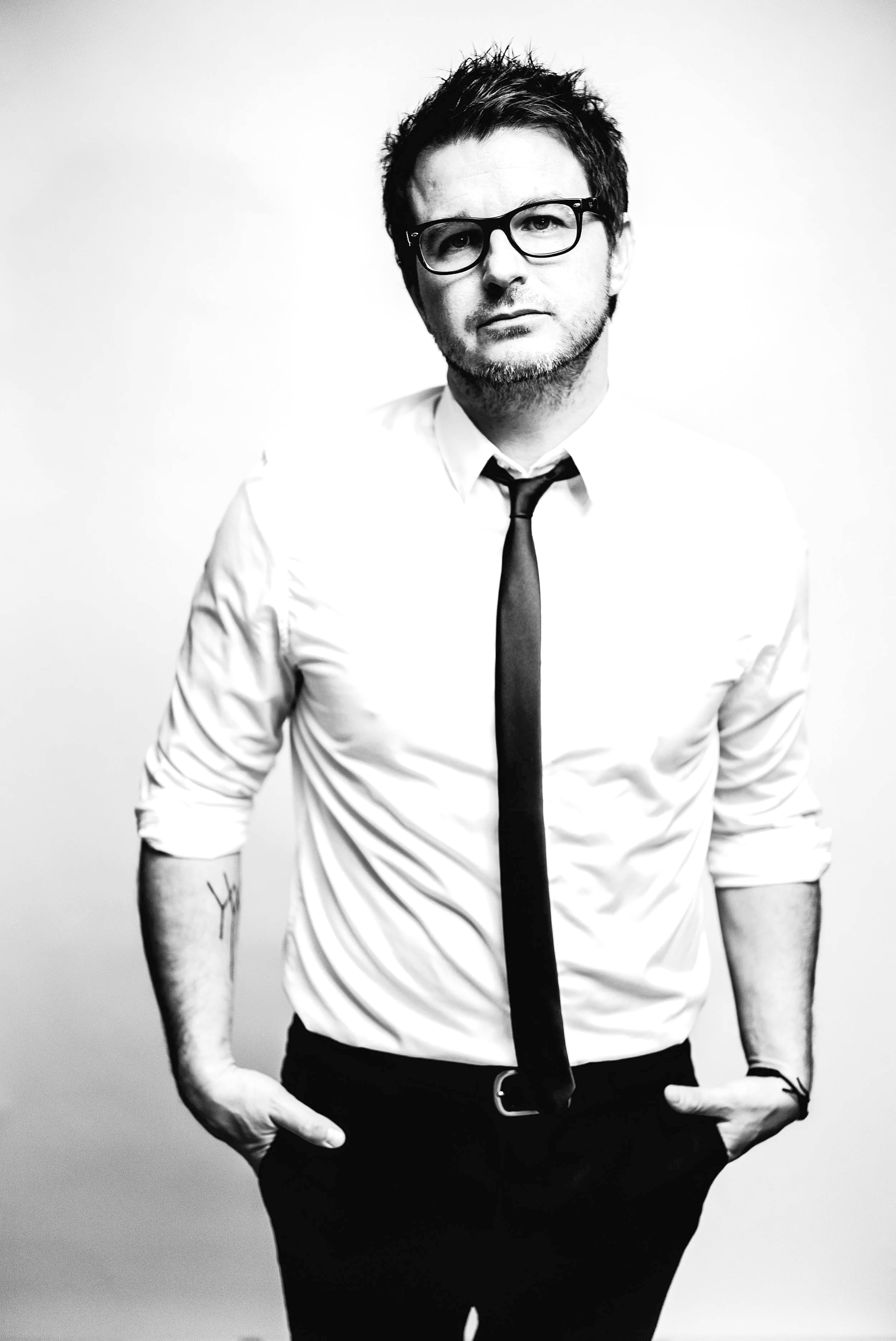 Black-and-white portrait of a man wearing glasses, a white shirt with rolled sleeves, a tie, and black pants, standing against a plain background with hands in pockets.