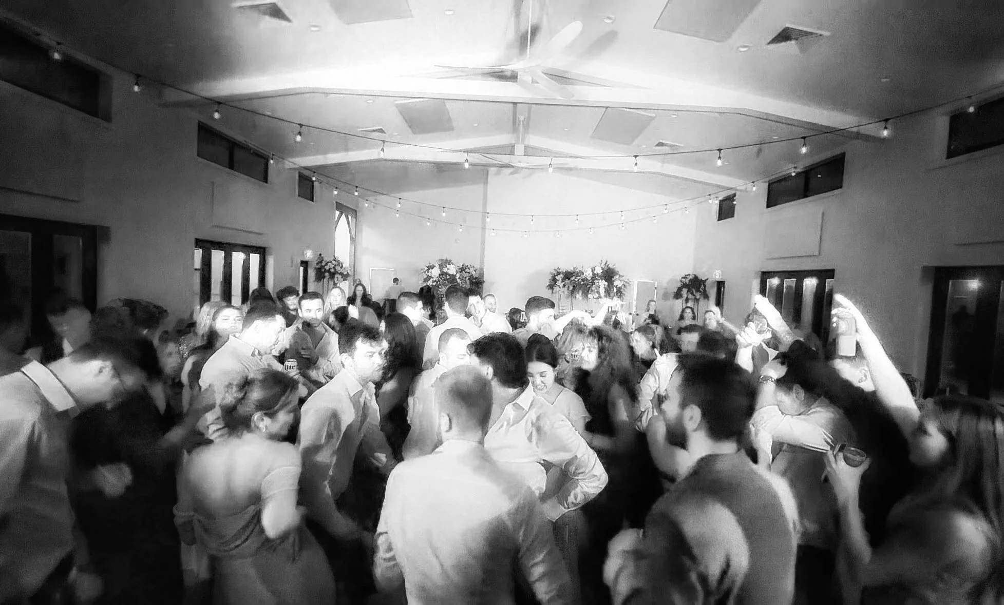 Black and white photo of a crowded dance floor at an indoor party or wedding reception, with people dancing and socializing, decorated with string lights and floral arrangements in the background.