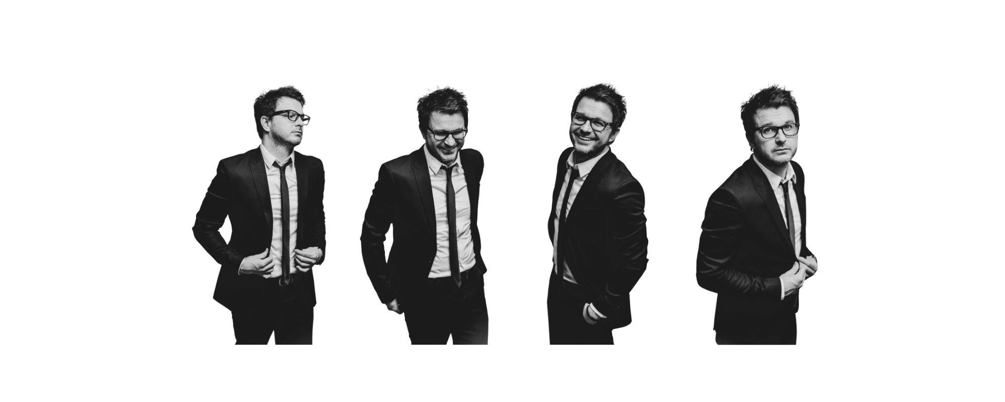 Four men in black suits and ties, with glasses, standing against a white background. The first man on the left is looking to the side with his hands in his pockets. The second man is looking down, smiling. The third man is smiling widely with his hands in his pockets. The fourth man is looking straight at the camera with a serious expression, holding his jacket with his hands.