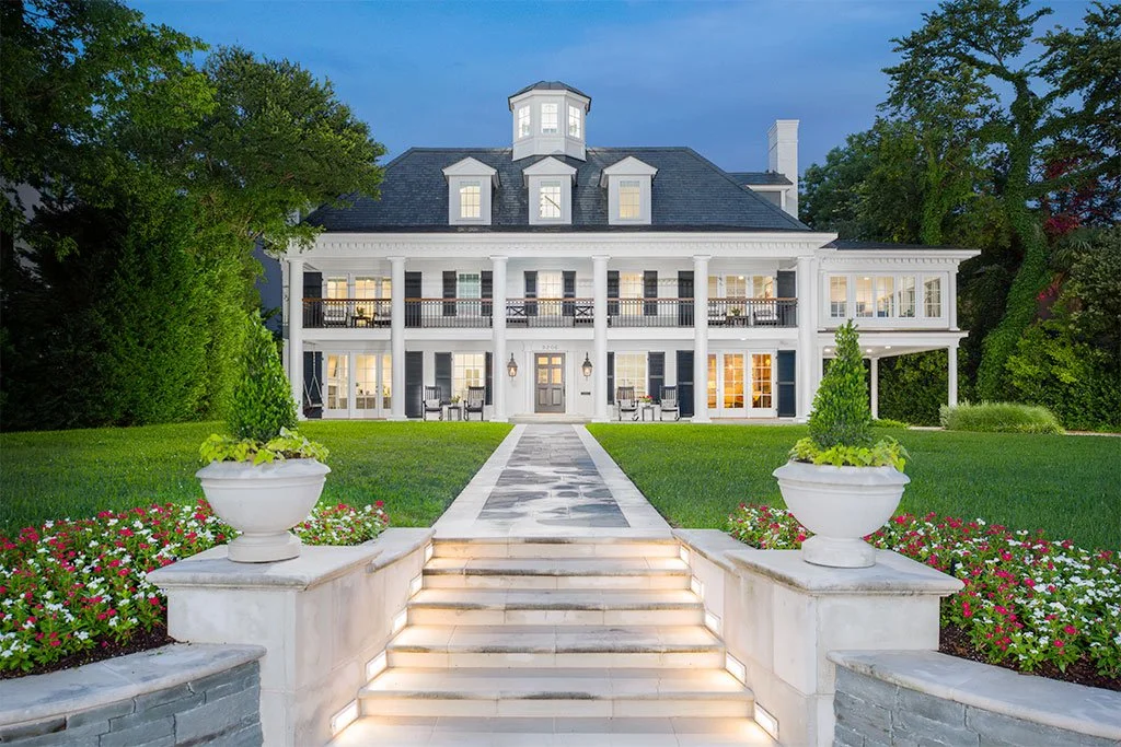 A large, elegant white house with a black roof, multiple windows, a balcony, and a prominent front door, surrounded by a well-maintained lawn, flower beds, and trees at dusk.