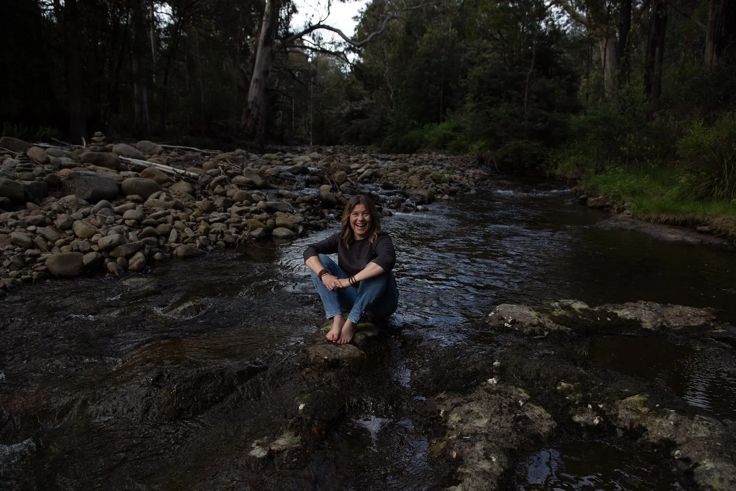 Kate Holman smiling on creek rock in forest in Tasmania | holistic wellness, nature connection, grounding wellbeing