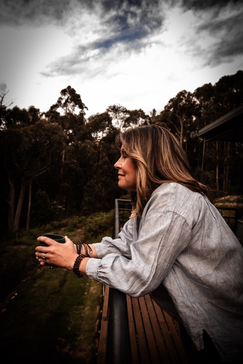 Kate Holman Naturopath holding cup on forest balcony in Tasmania | peaceful wellbeing lifestyle, holistic health illustration