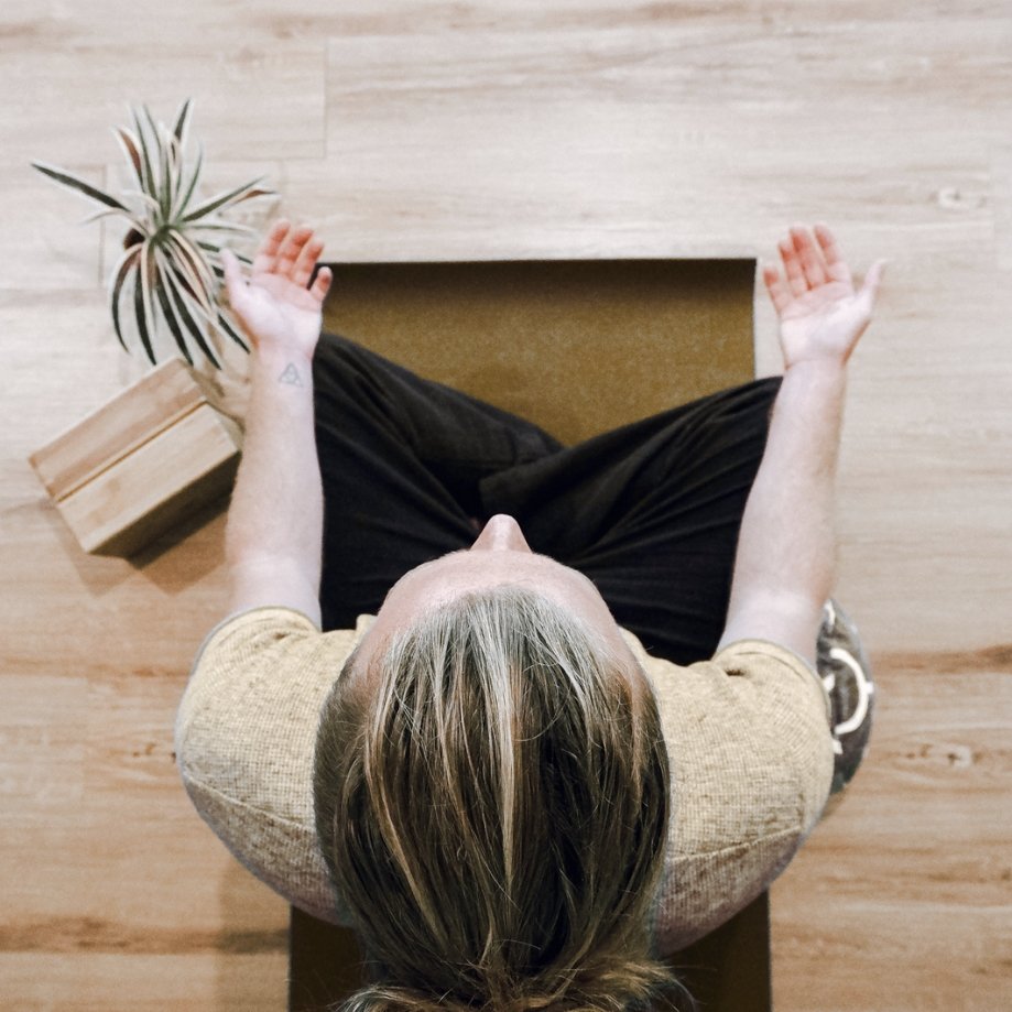 A person sitting cross-legged on a yoga mat in a room with wooden flooring, palm plant, and wooden blocks, practicing meditation or mindfulness.