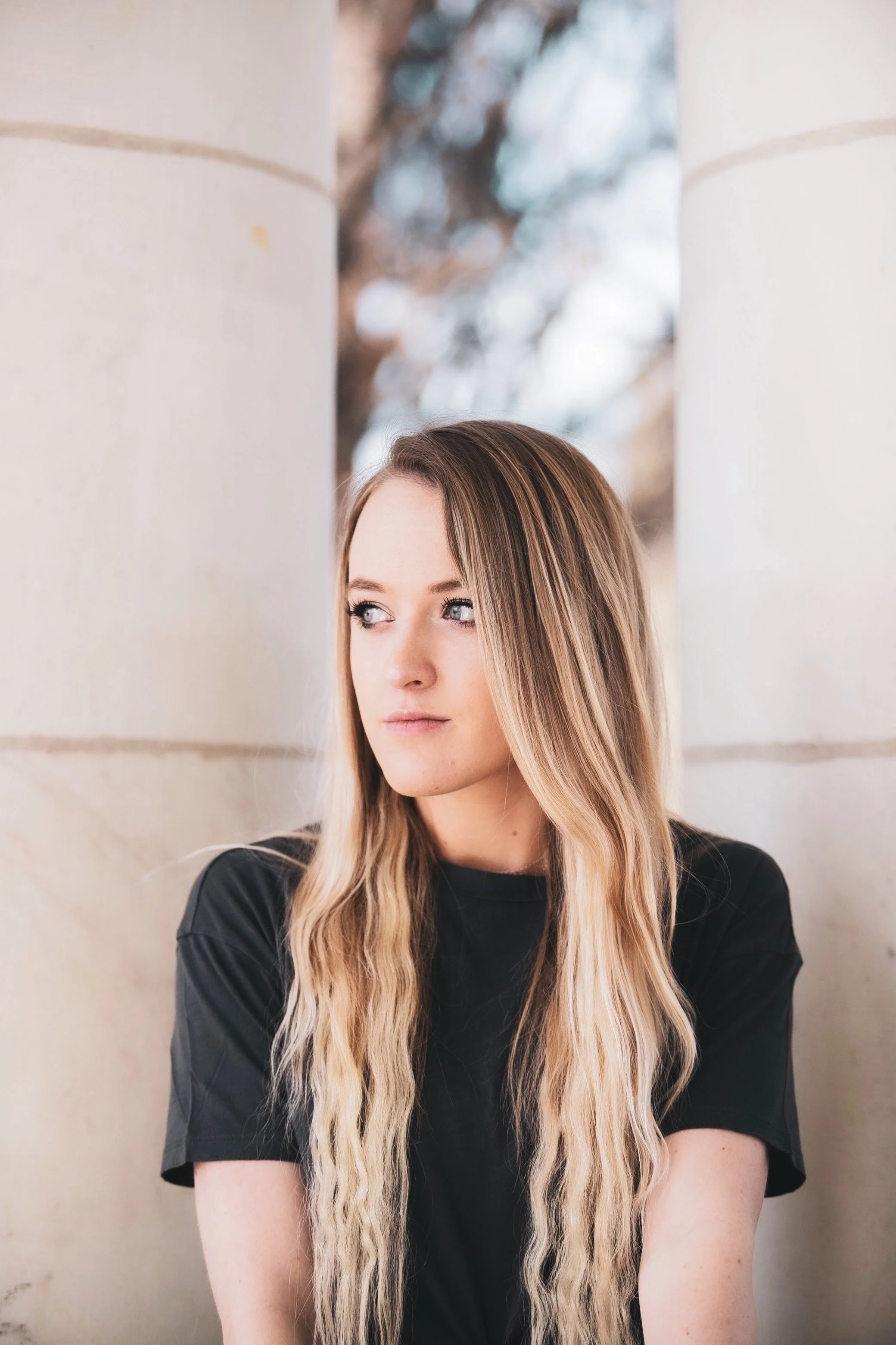 A young woman with long, wavy blonde hair and blue eyes looking to her left while standing against a stone wall with trees in the background.