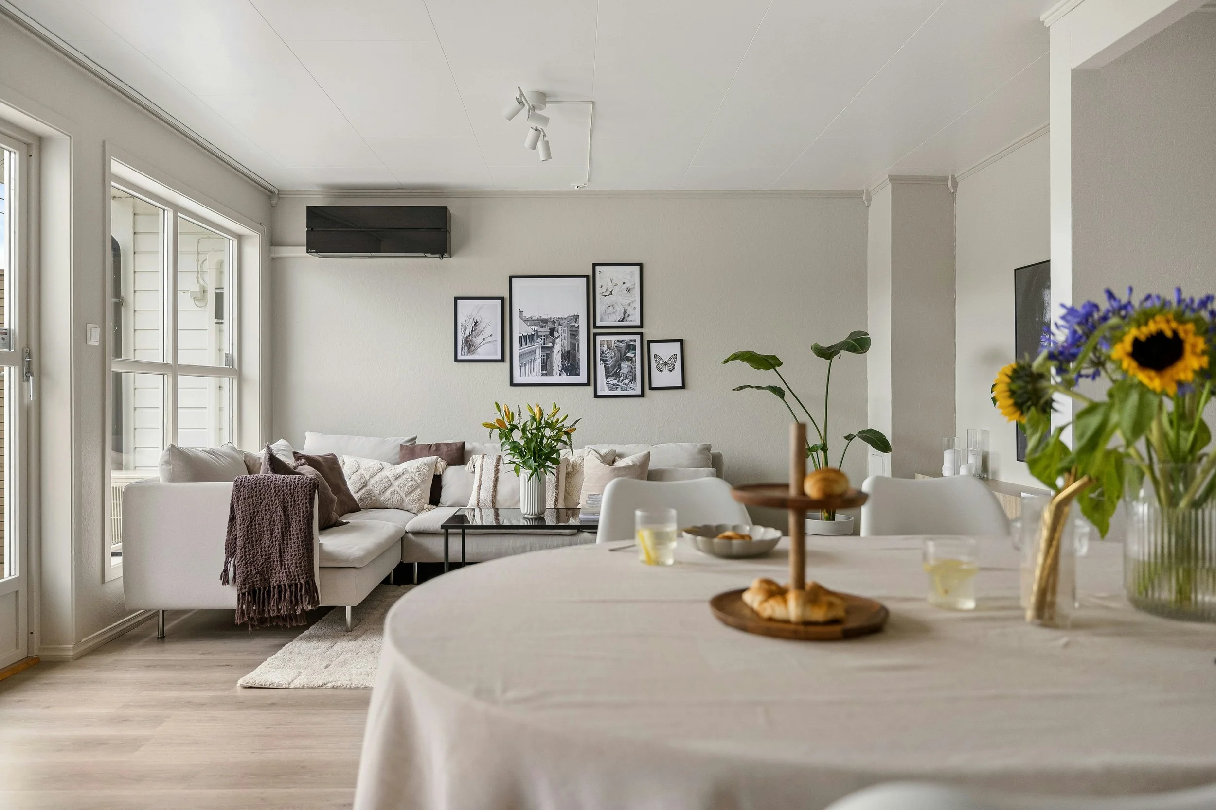 A bright living room with a white sectional sofa, framed black-and-white artwork on the wall, and a dining table with flowers and snacks in the foreground.