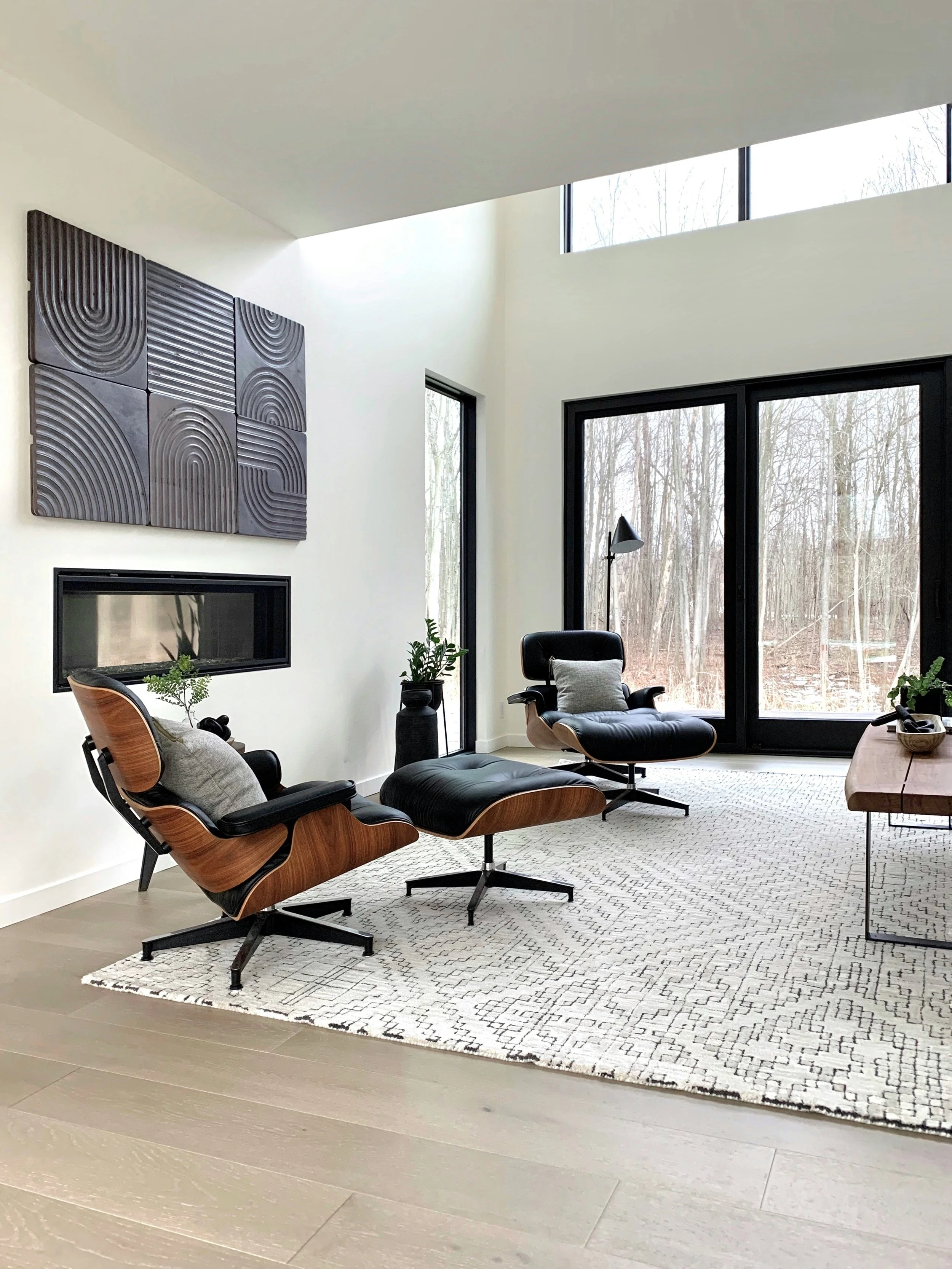 Modern living room with white walls, large windows, a fireplace, and contemporary furniture including two black leather chairs and an ottoman on a patterned rug.