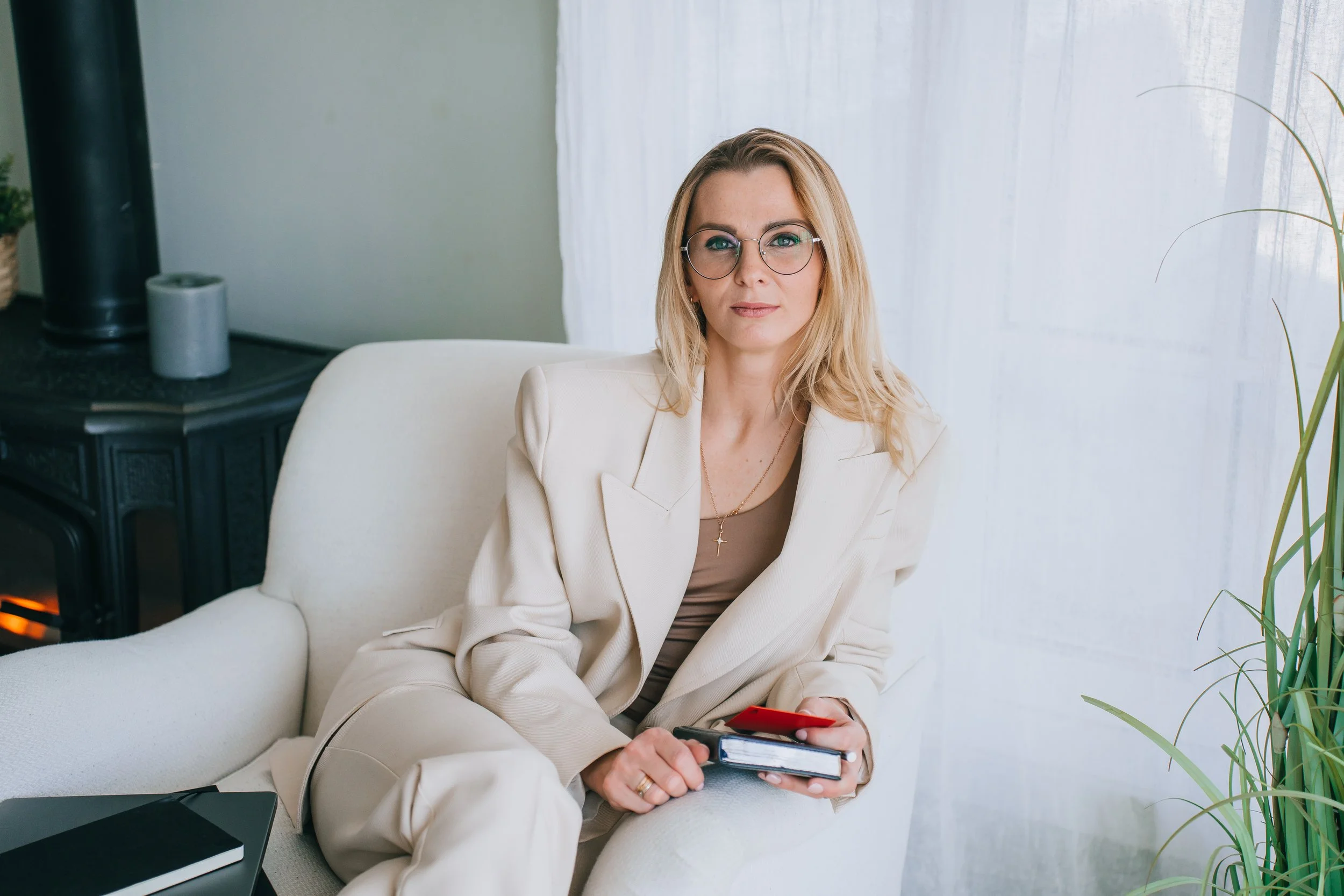 A woman with blonde hair, glasses, wearing a cream-colored suit, sitting on a white armchair, holding a smartphone and a notebook, in a brightly lit room with large white curtains and green plants.