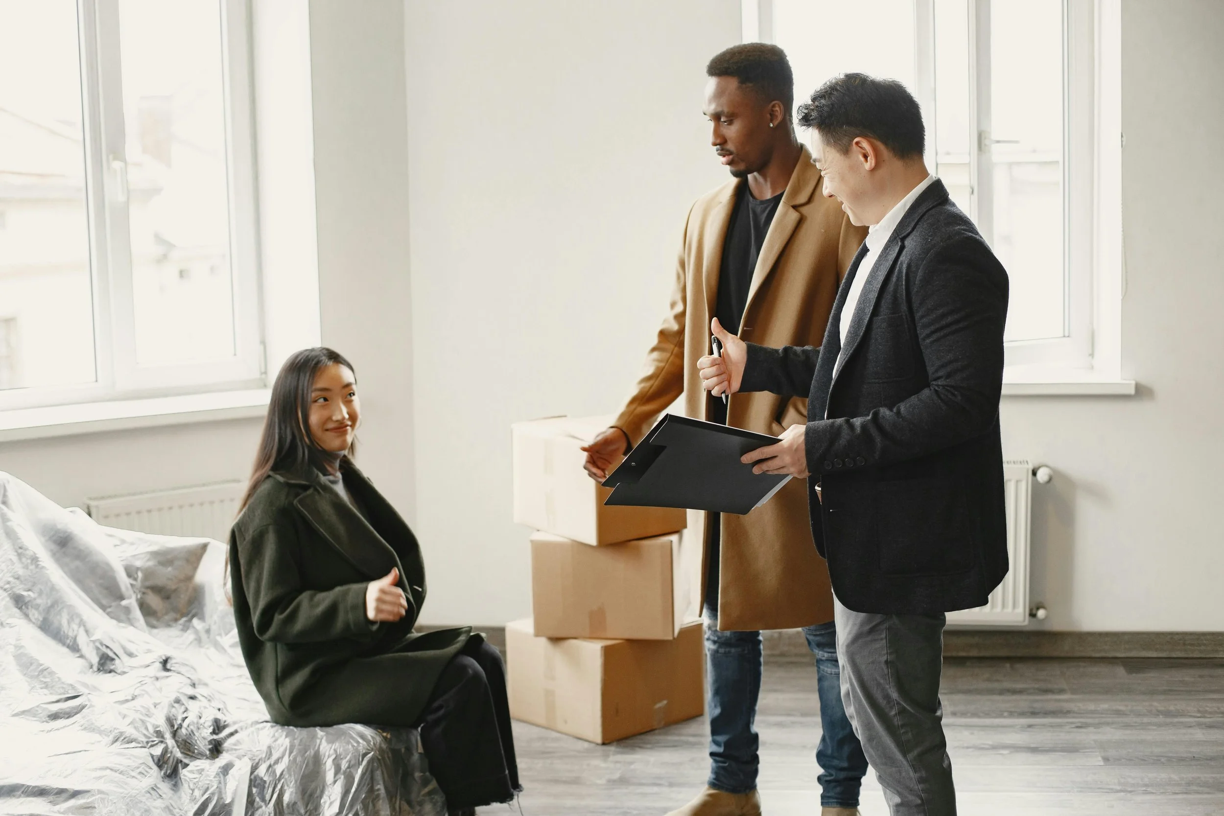 A young woman sitting on a bed in a room with stacked boxes, talking to two men standing nearby, one holding a clipboard, in a casual office or apartment setting.