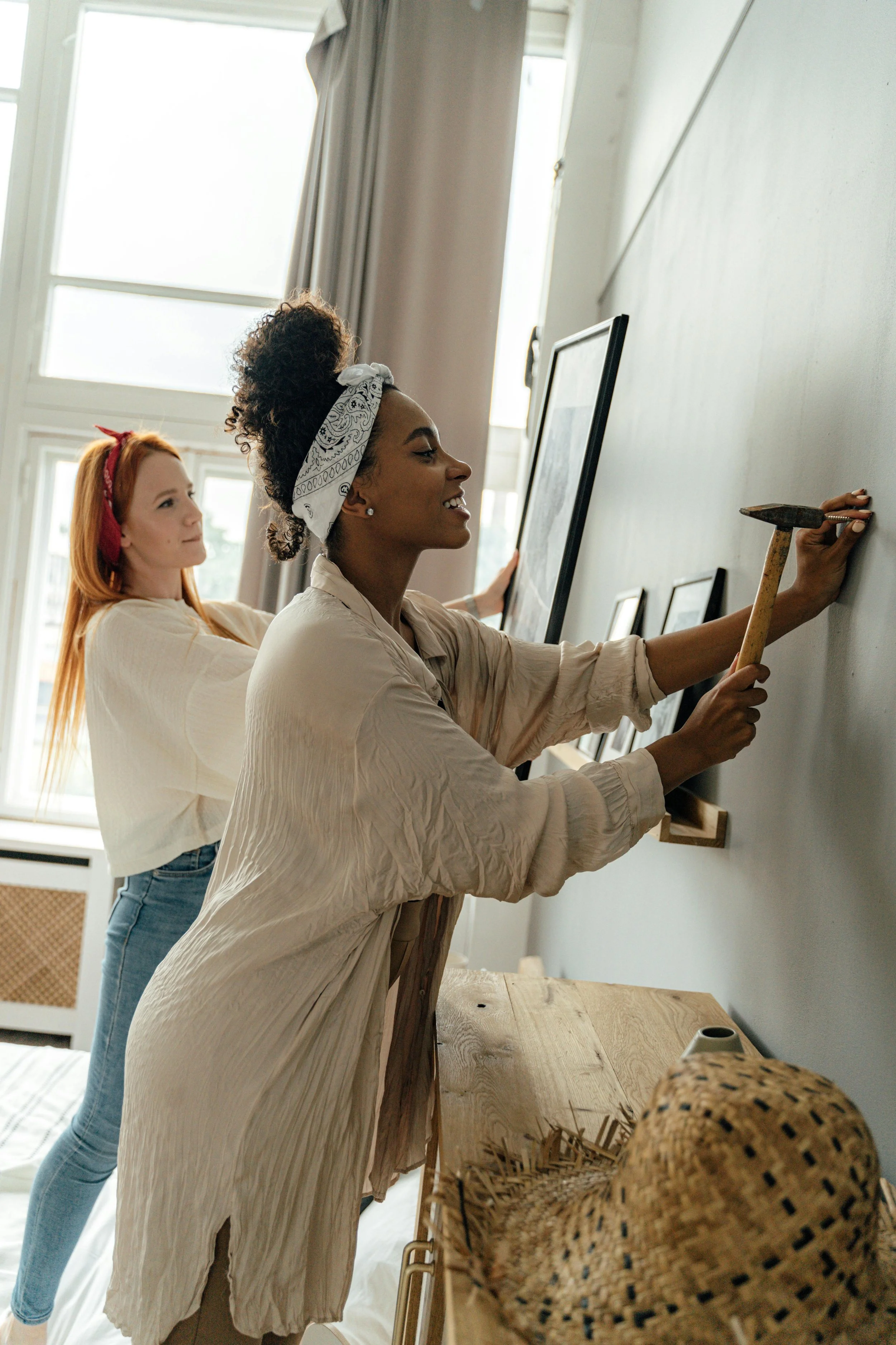 Two women hanging picture frames on a gray wall in a well-lit room.