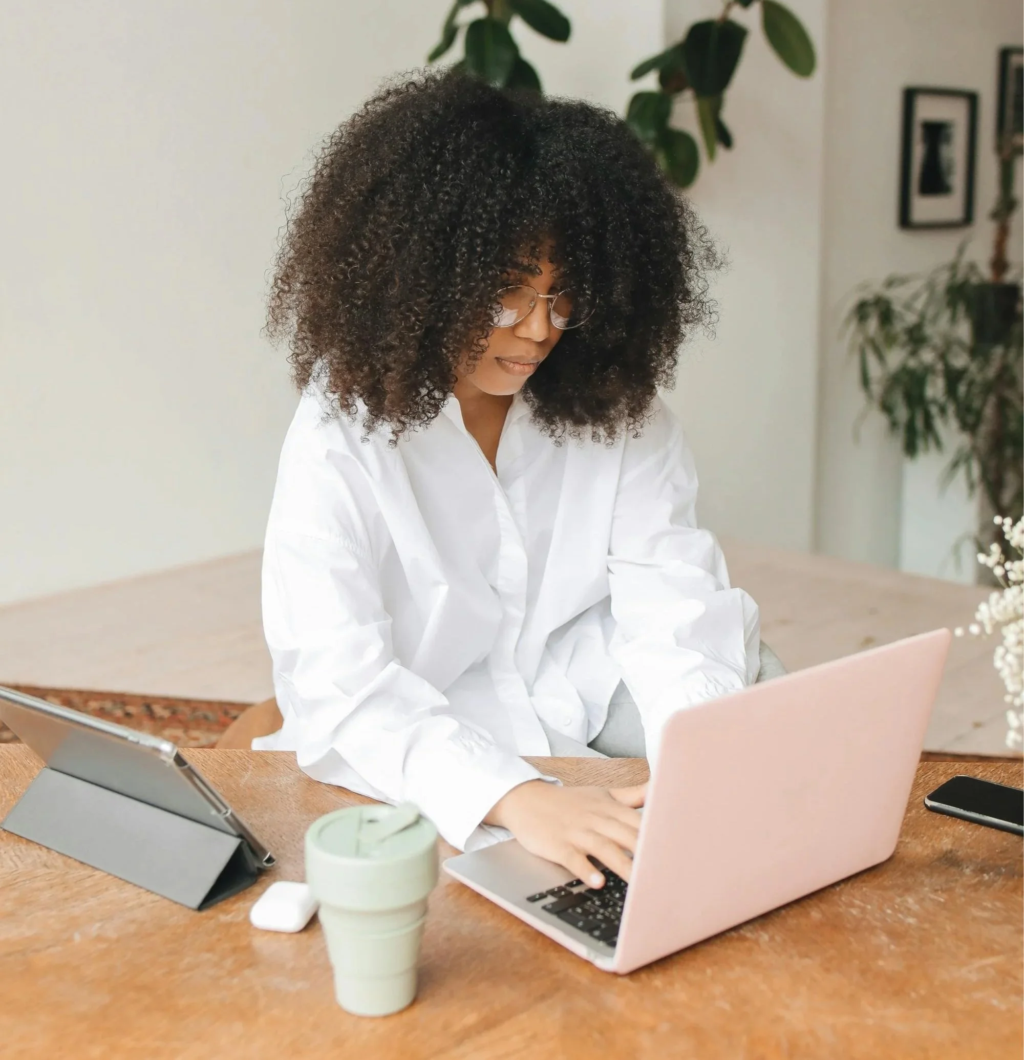 A woman with curly hair and glasses working on a pink laptop at a wooden table, surrounded by a tablet, coffee cup, phone, and potted plant in a bright room.