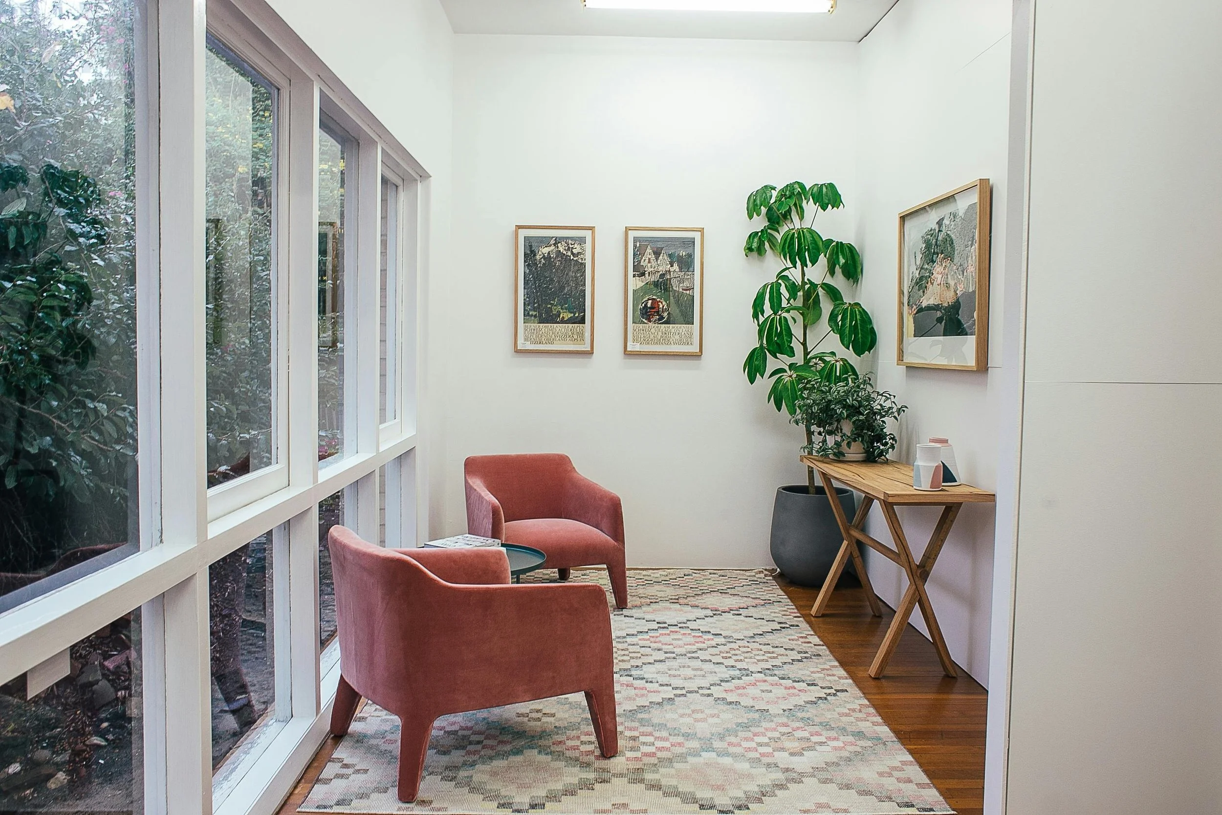 Cozy sitting area with two pink upholstered armchairs, a small black side table, a large potted green plant, framed artwork on white walls, a wooden console table with decor, a window with greenery outside, and a patterned rug on hardwood floor.