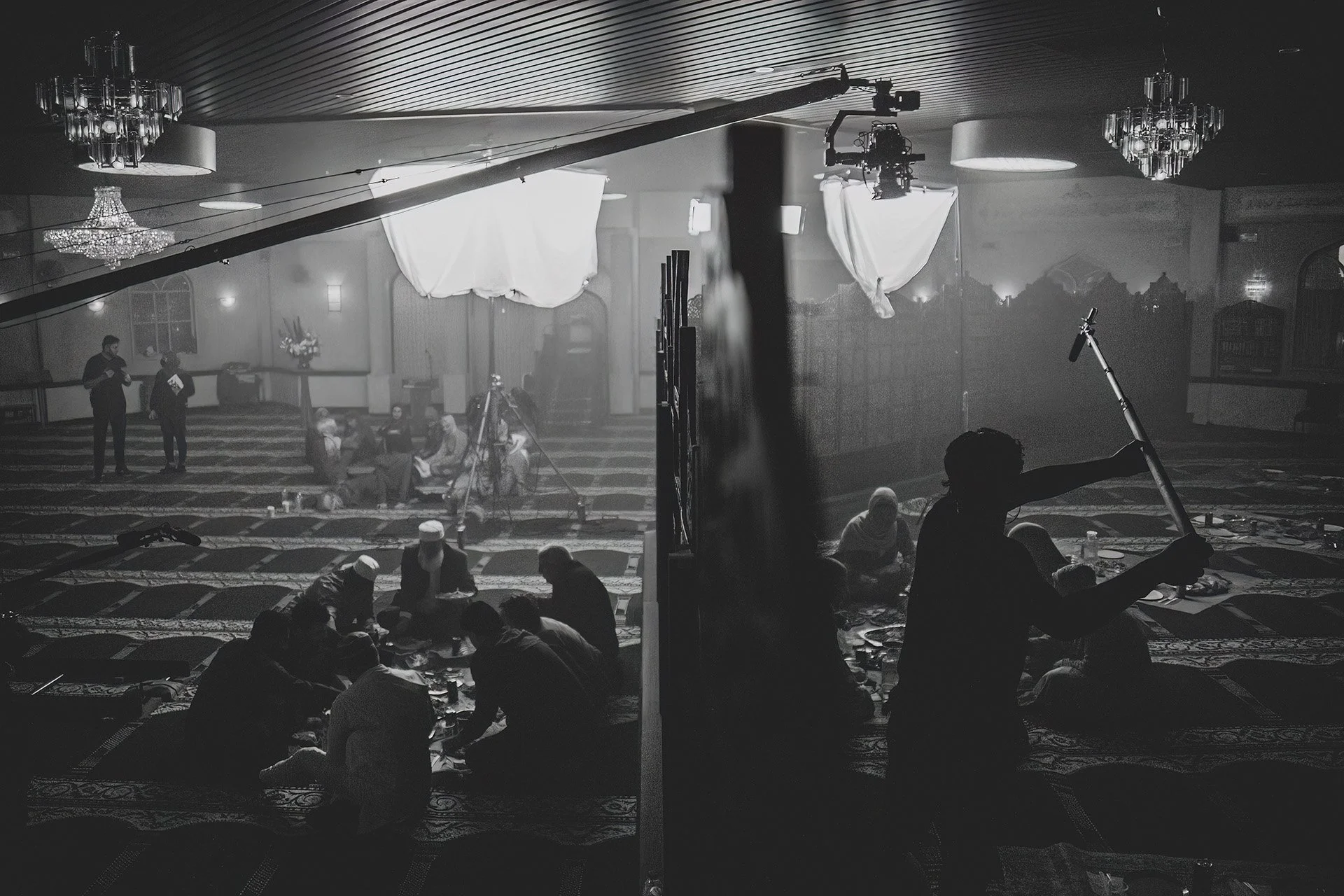 People in a mosque with prayer rugs, some sitting and some standing, during filming of a religious event with lighting and camera equipment.