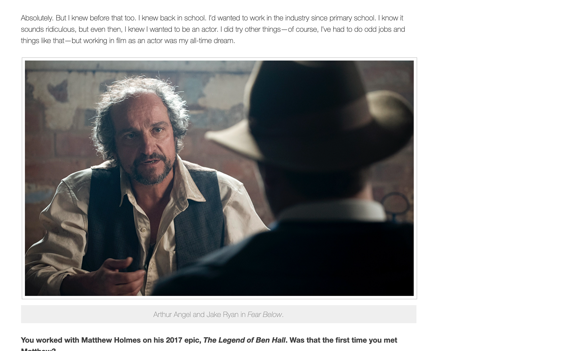 Two men having a serious conversation in a dimly lit room with brick walls. One man with curly gray hair and a beard, wearing a vest, looks intently at the other man, who is wearing a wide-brimmed hat.