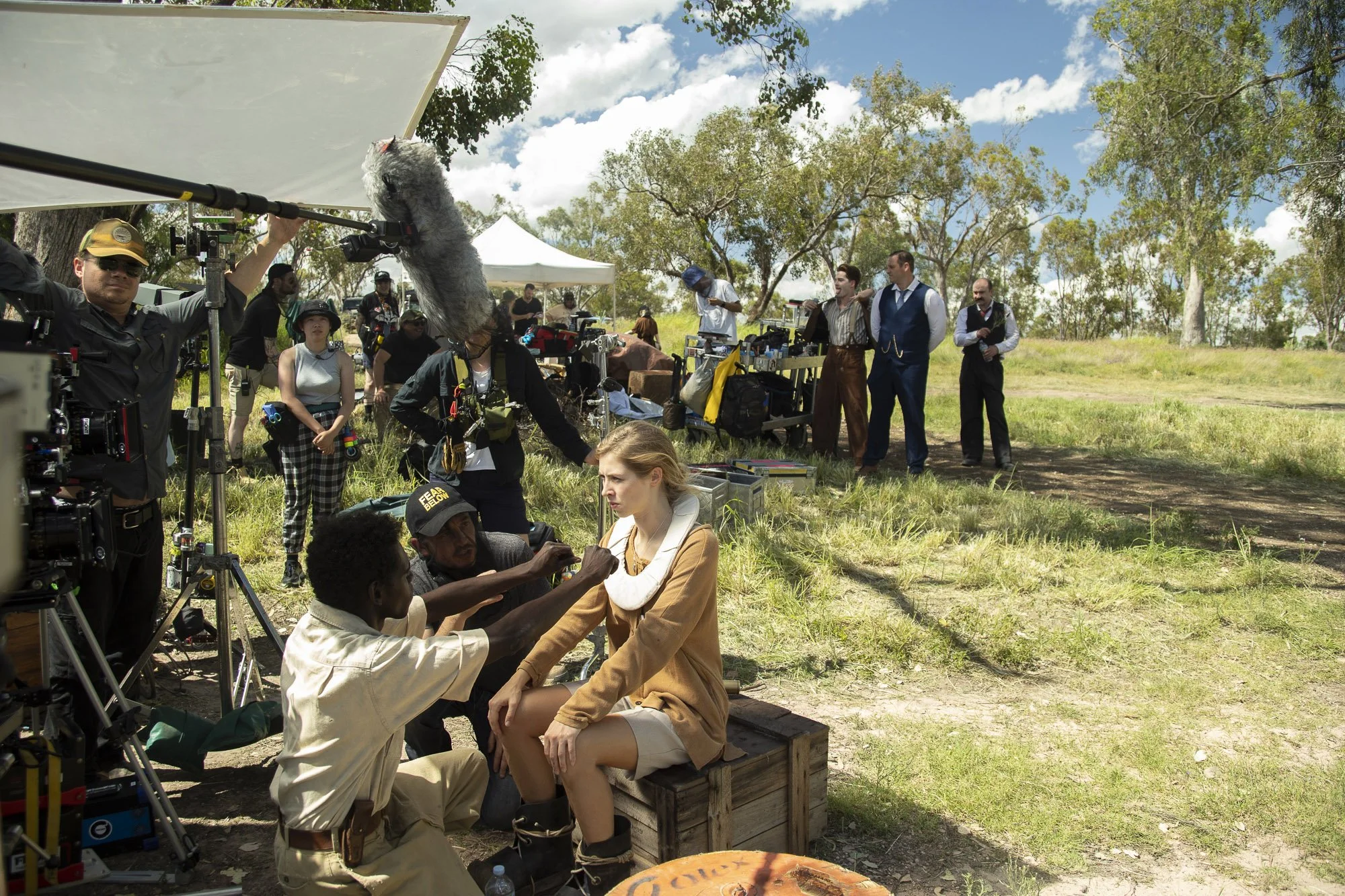Film crew filming outdoor scene with young woman seated on wooden crate, surrounded by equipment, crew members, and actors in period costumes, under a partly cloudy sky with trees and grassy landscape in background.