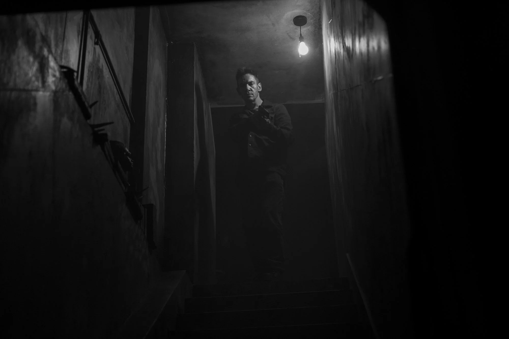 A man standing in a dimly lit narrow corridor or staircase, facing the camera with a serious expression, with a light bulb hanging from the ceiling.