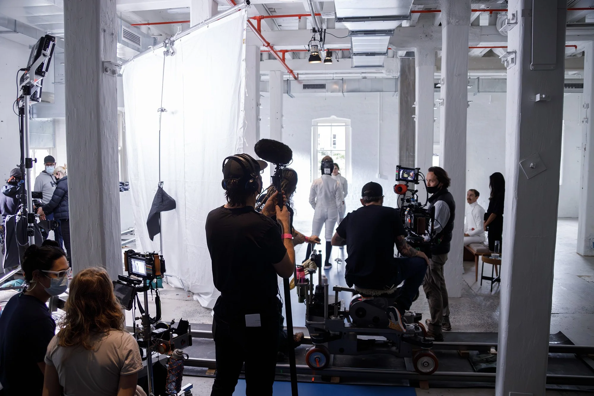 Film crew filming scene in a white studio with white walls and large window, including camera operators, director, and actors, with lighting and microphone equipment.