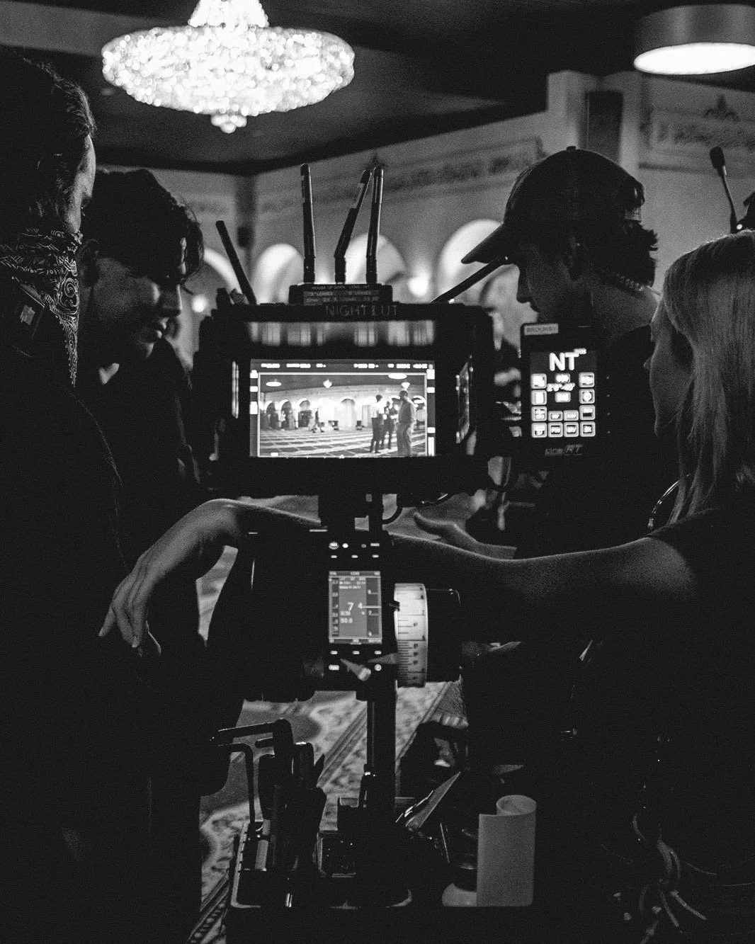 Black and white image of a film camera and crew members on a movie set, with people working behind the scenes in a dimly lit room featuring a chandelier and arched windows.