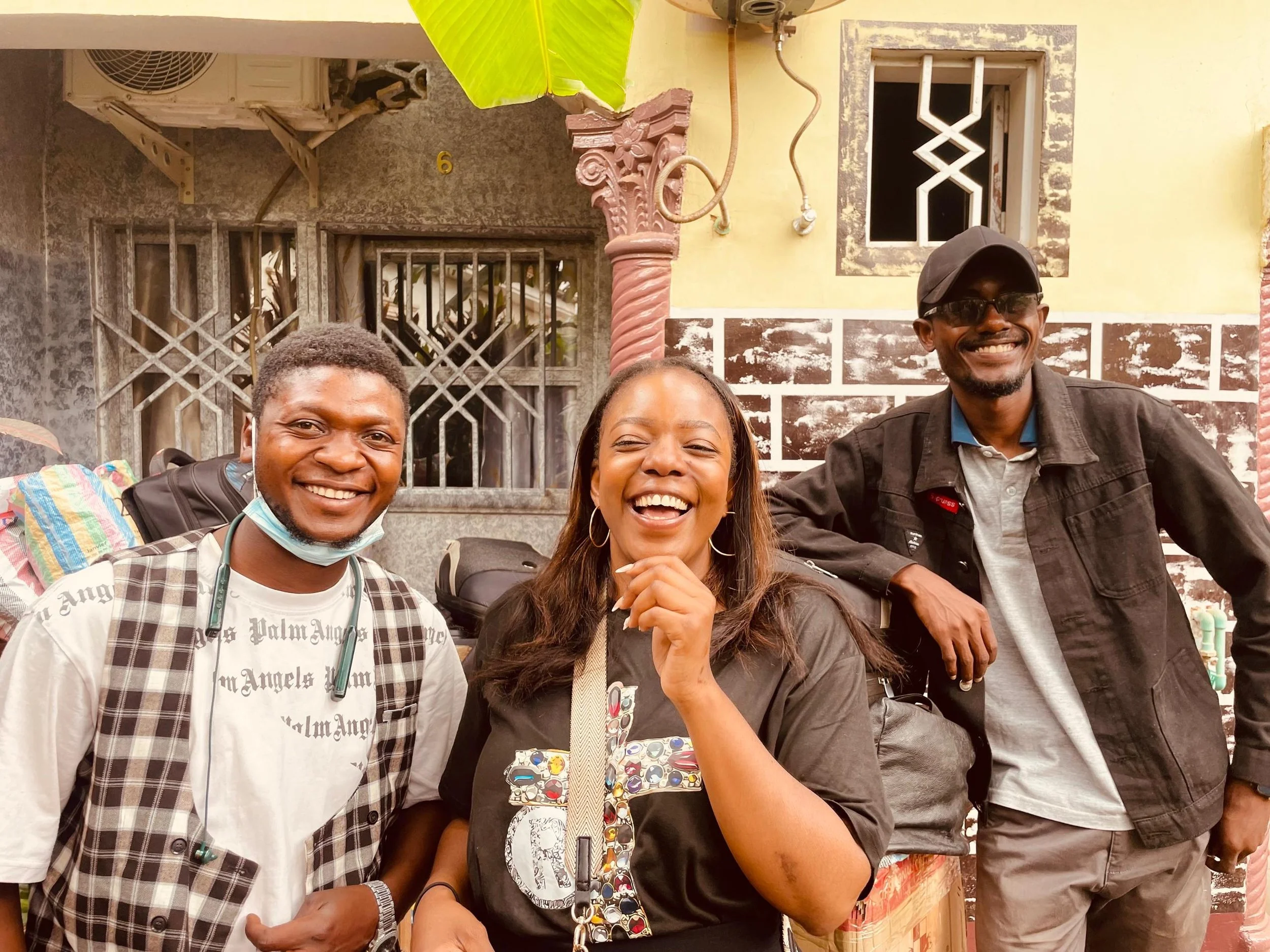 Three smiling people standing outdoors in front of a building with windows and brick wall, one wearing a mask, two without masks, laughing and posing for the photo.