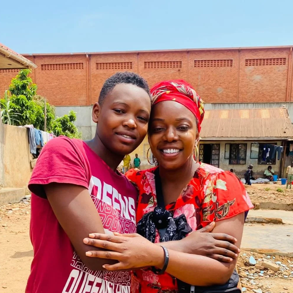 A woman and a girl hugging and smiling outdoors in front of a brick wall and a small building. The woman is wearing a red headscarf and a red floral dress, the girl is wearing a red T-shirt.