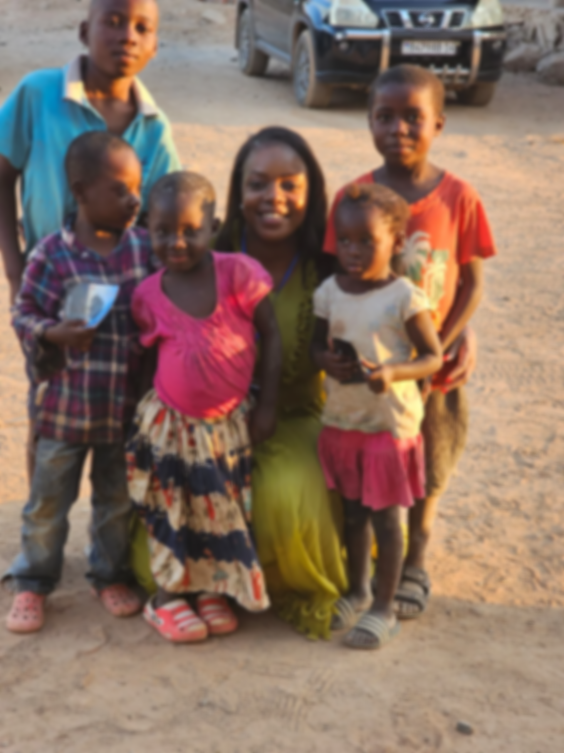 Group of six children and one woman standing on a dirt ground, with a police vehicle in the background.