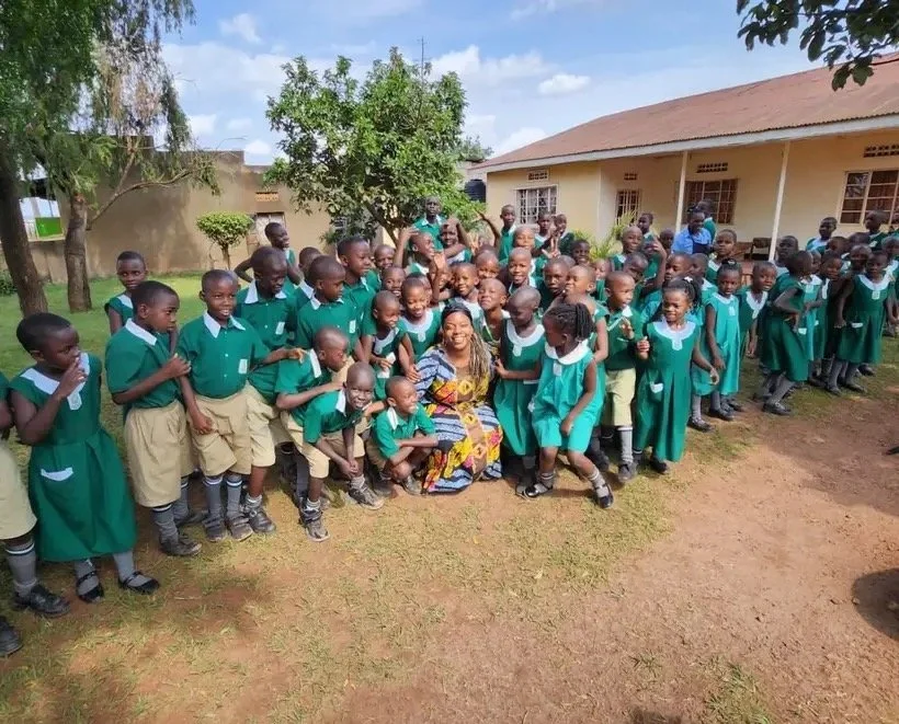 A large group of school children in green uniforms gathered outdoors around a woman in a colorful dress, with a school building and trees in the background.