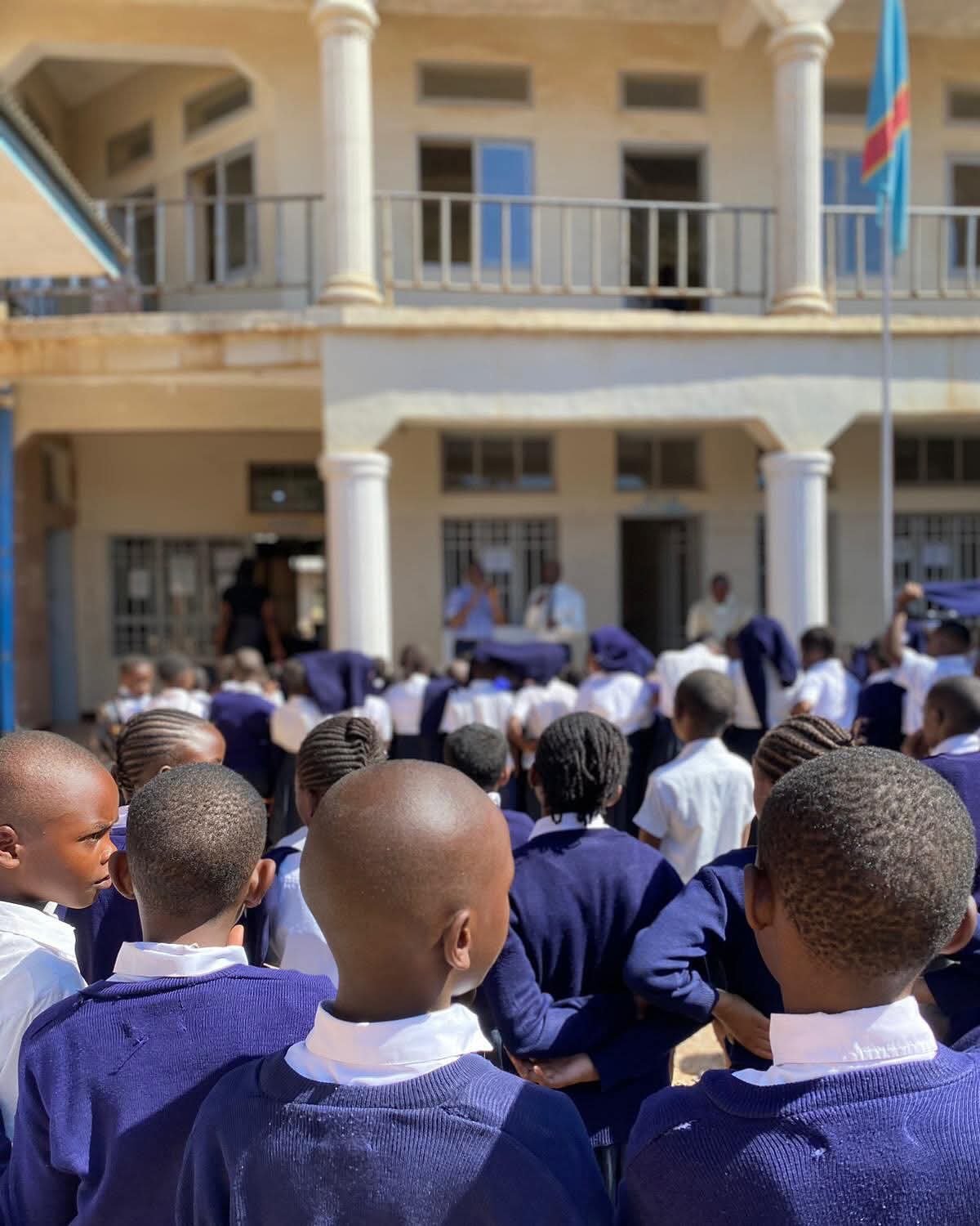 A group of students in school uniforms gathered outside a building, facing a stage where three people are speaking.
