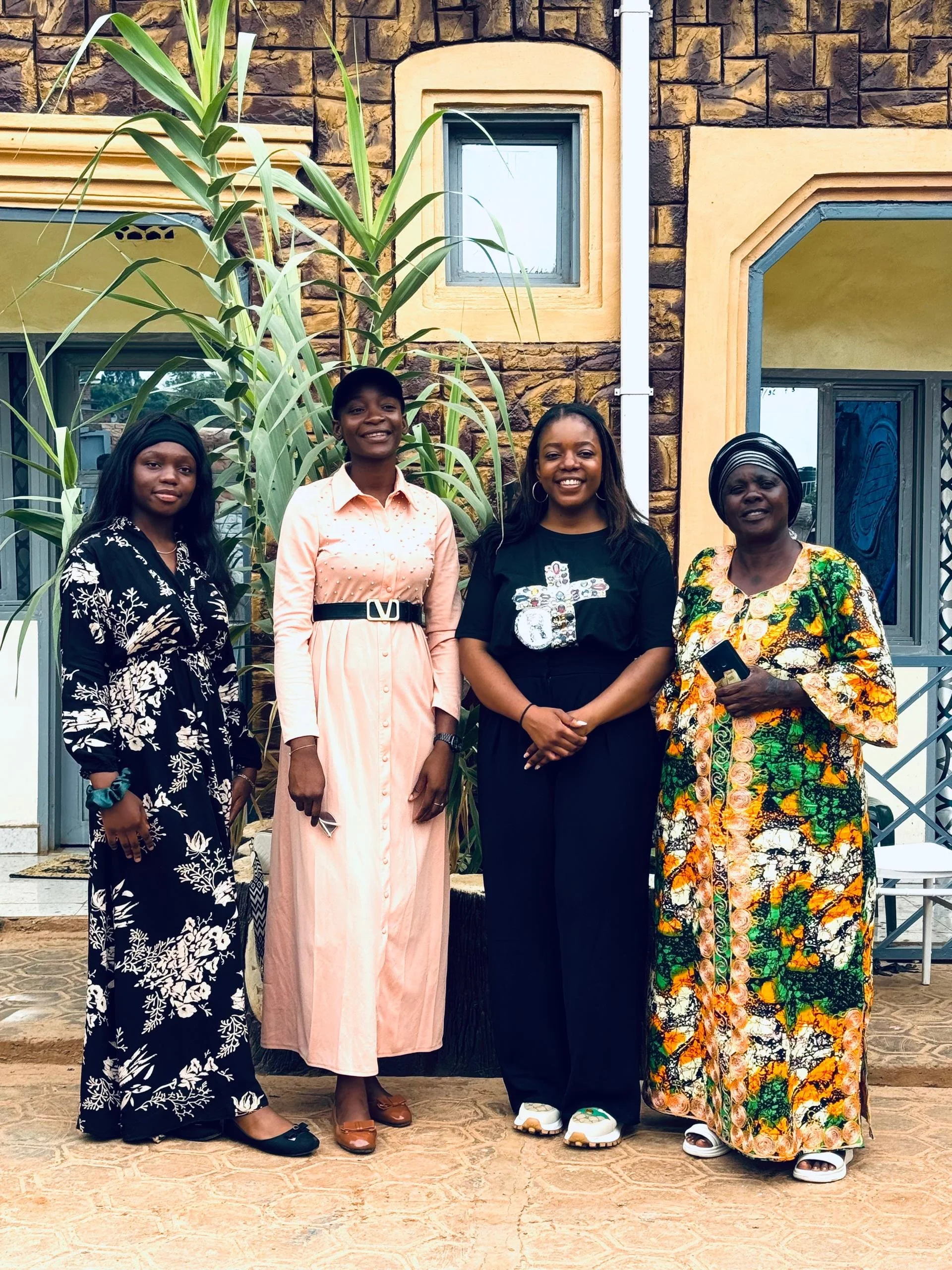 Four women standing outdoors in front of a brick building with windows and a green plant. They are smiling and dressed in different styles of clothing.