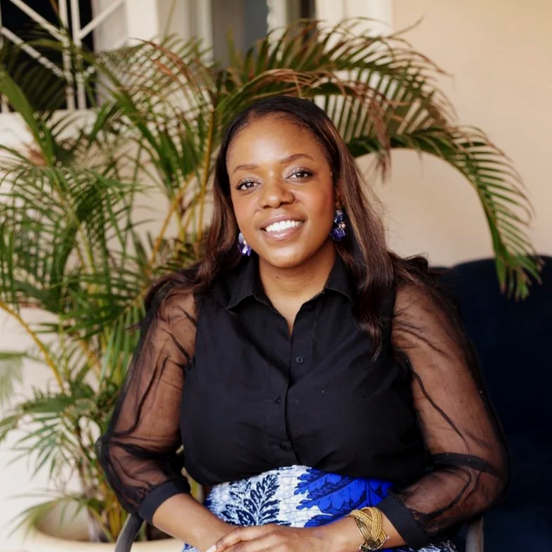 A woman with long dark hair, wearing a black top with sheer sleeves and blue patterned skirt, smiling while sitting indoors with green plants in the background.