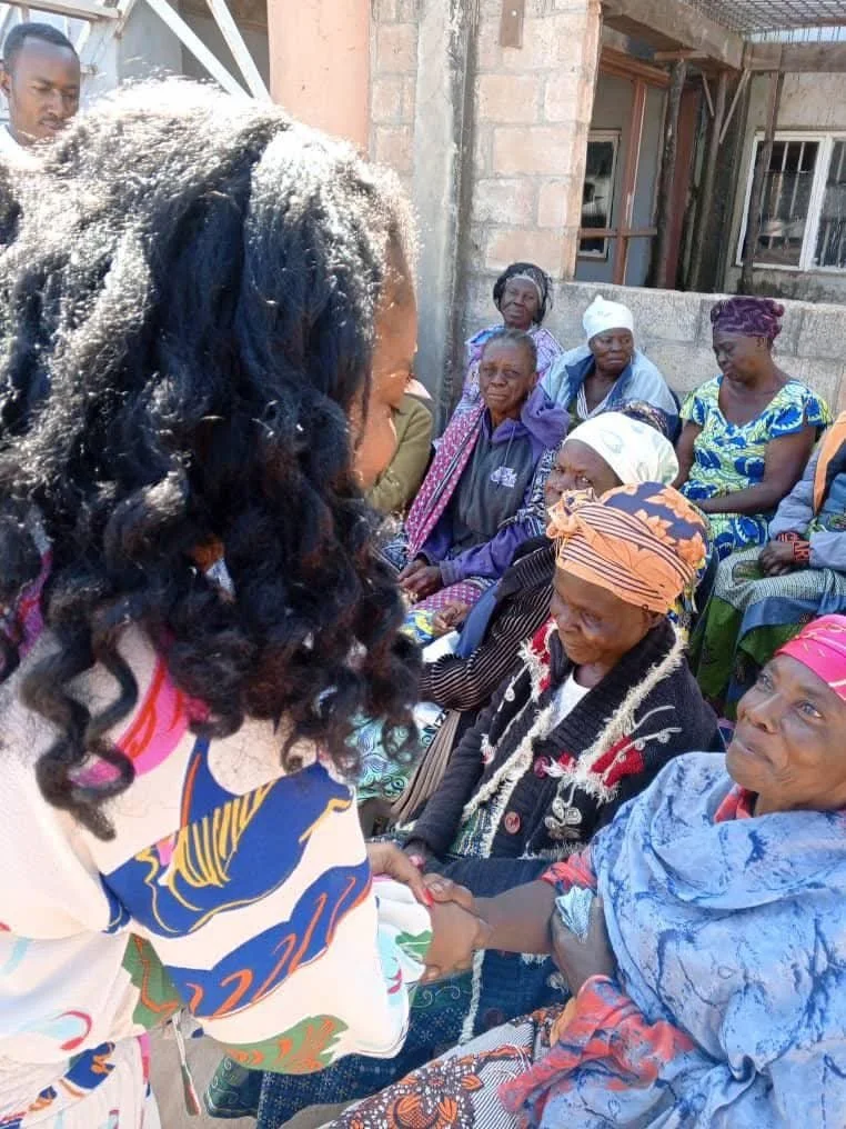 A woman in the foreground shaking hands with an elderly woman, while a group of women sit and stand around them outside of a building, engaging in a gathering.