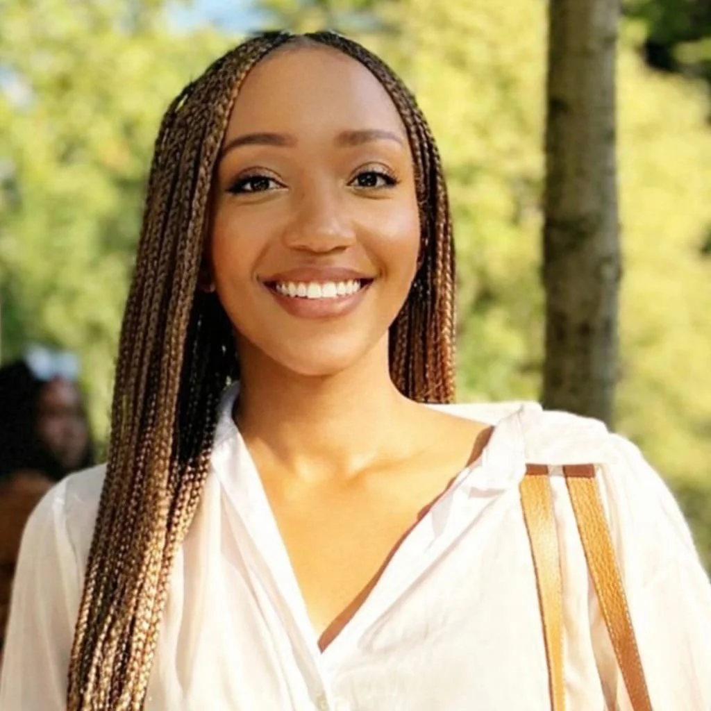 Smiling woman with long braided hair outdoors in a park, wearing a white blouse and carrying a brown shoulder bag.