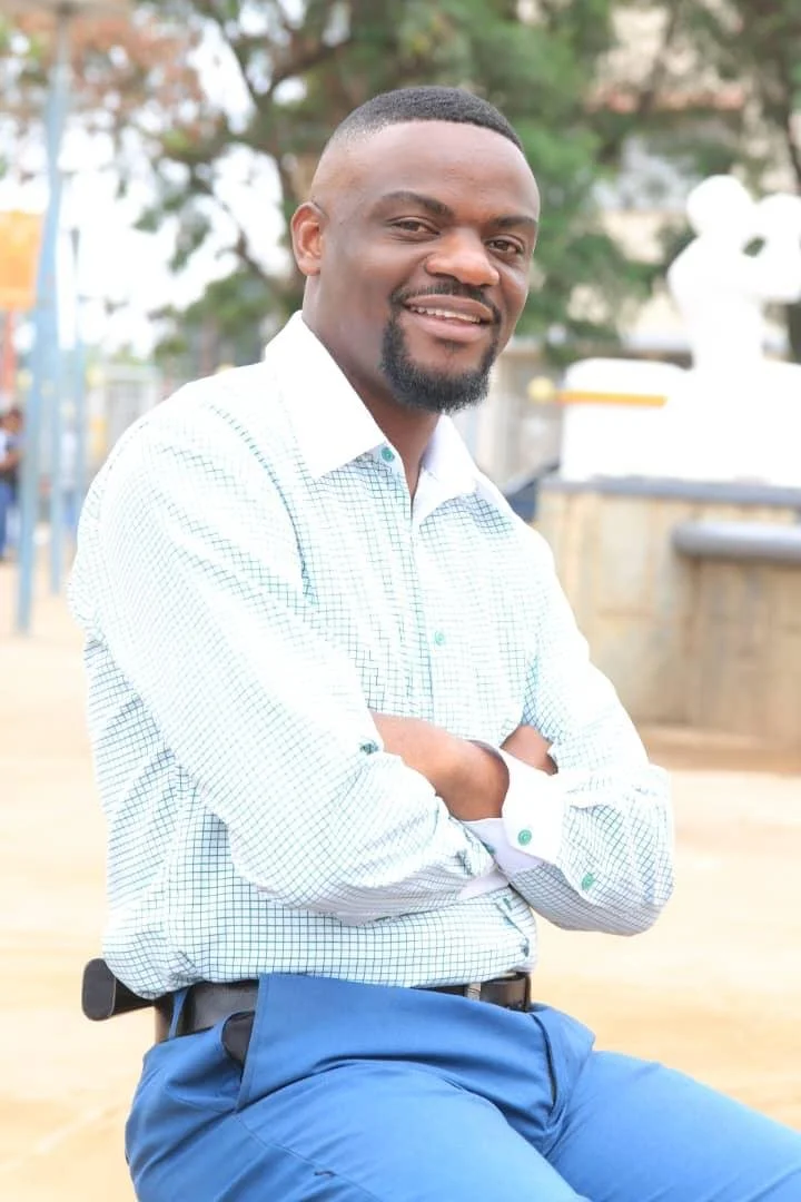 A man with short black hair and a beard smiling at the camera, wearing a white checkered shirt and blue pants, standing outdoors with a blurred background of trees and structures.