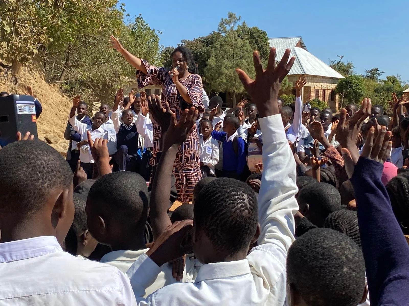 A woman speaking into a microphone and gesturing with her hand, standing on an elevated platform or seat, surrounded by a crowd of students raising their hands outdoors during the day.