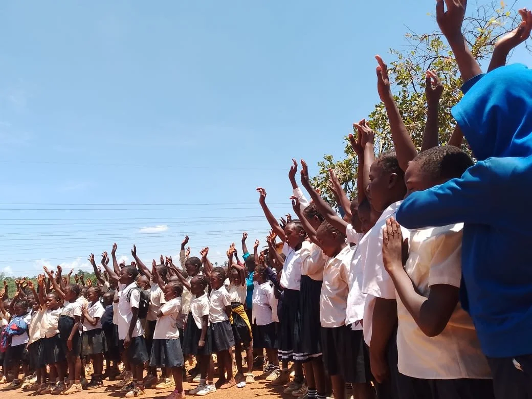 A group of children in school uniforms standing outdoors under a clear blue sky, raising their hands in the air.