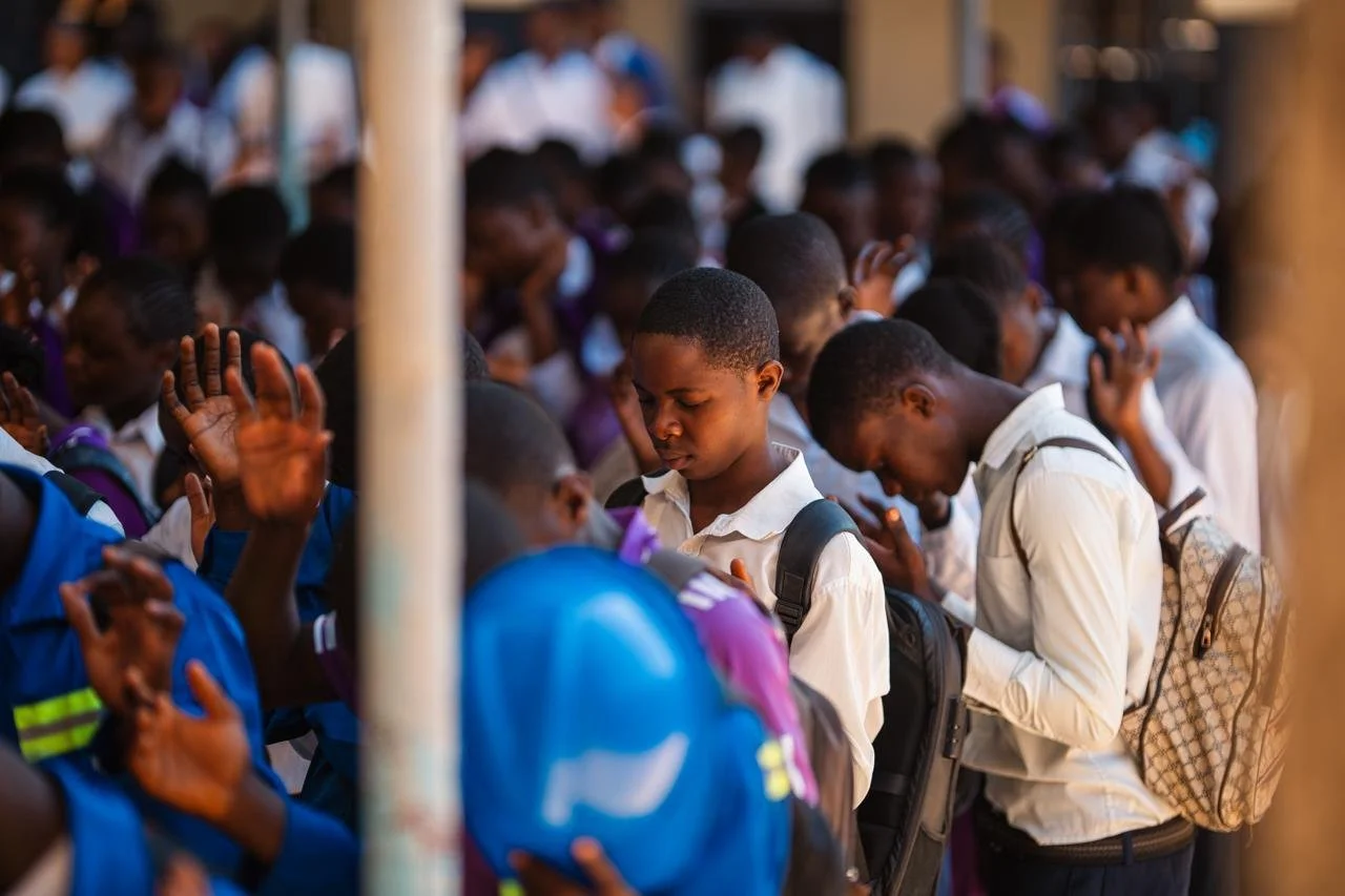 Schoolchildren praying with heads bowed and eyes closed, some with hands raised, wearing uniforms and backpacks.