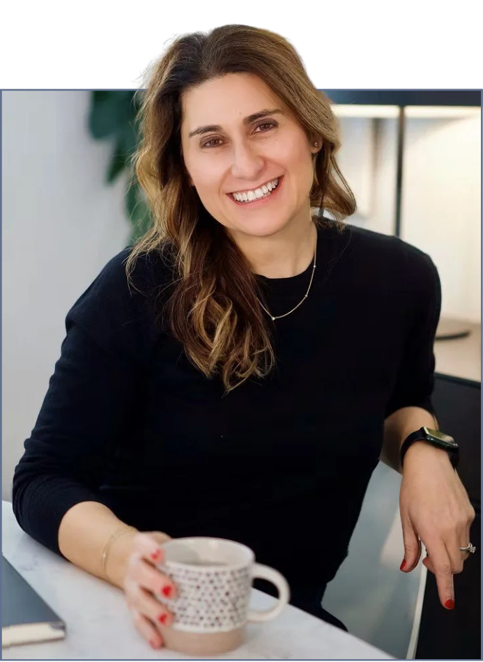 A woman with brown wavy hair smiling while sitting at a table with a mug in her hand, wearing a black top, gold jewelry, and a smartwatch, in a cozy indoor setting.