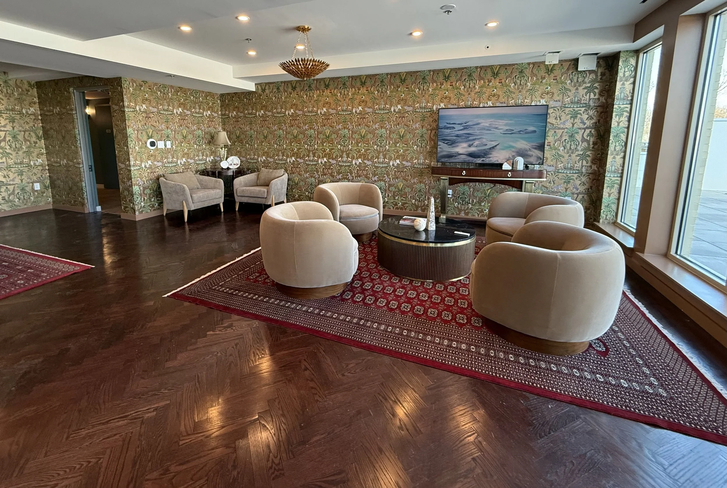 Luxurious hotel lounge with beige rounded chairs arranged around a black and gold coffee table on a red patterned rug, wooden floors, and tropical wallpapered walls. A wall-mounted flat-screen TV is above a wooden console, with large windows to the r