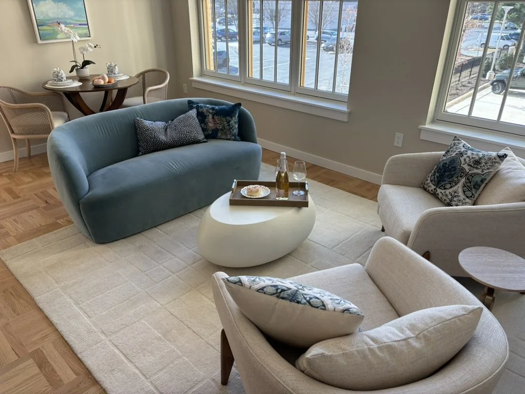 Living room with large windows, beige and blue furniture, a white oval coffee table with drinks, and a dining area with a round table and four chairs.