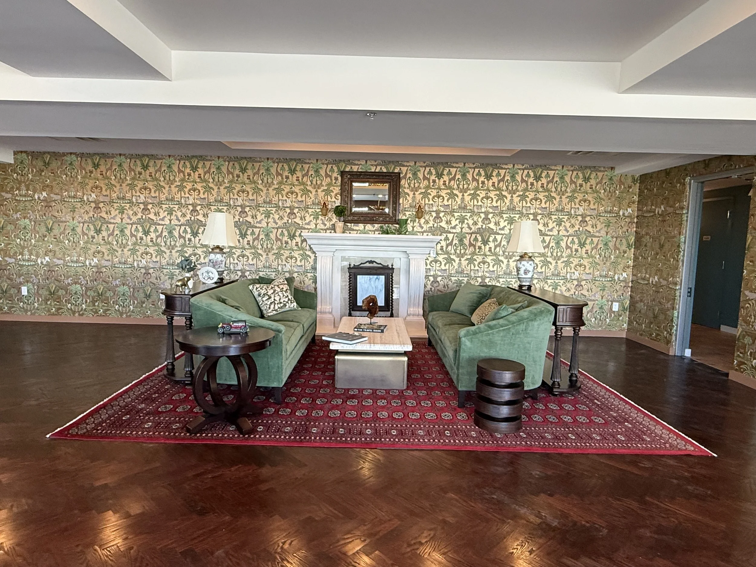 Living room with two green sofas facing each other, a fireplace with a mirror above, a red patterned rug, and side tables with lamps.