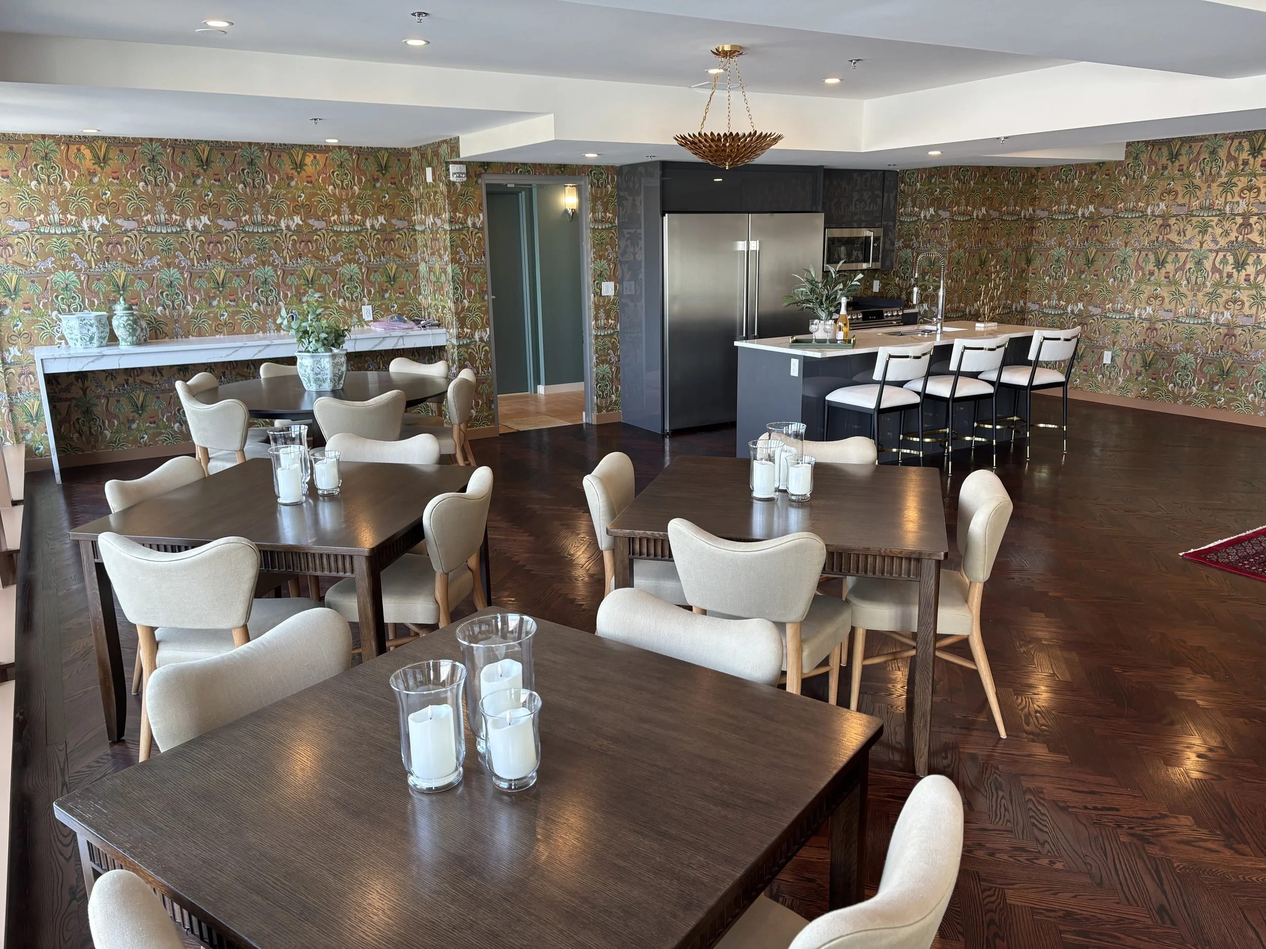 Interior of a modern dining and kitchen area with multiple wooden tables, beige upholstered chairs, candles, a colorful patterned wallpaper, and a kitchen island with barstools, stainless steel appliances, and decorative plants.