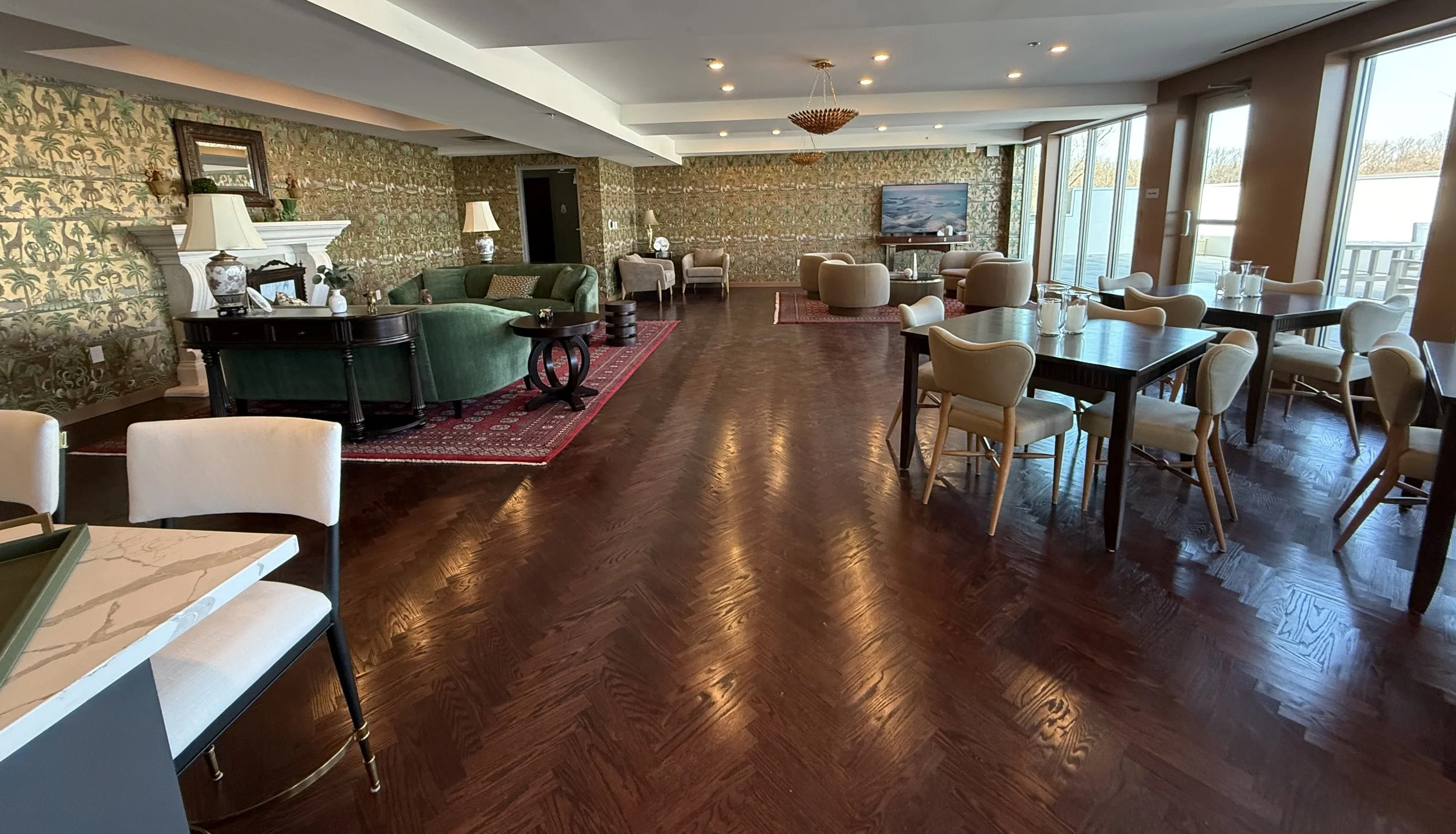 Spacious hotel lounge with wooden flooring, multiple arranged tables with chairs, green and beige couches, a TV, and large windows providing natural light.