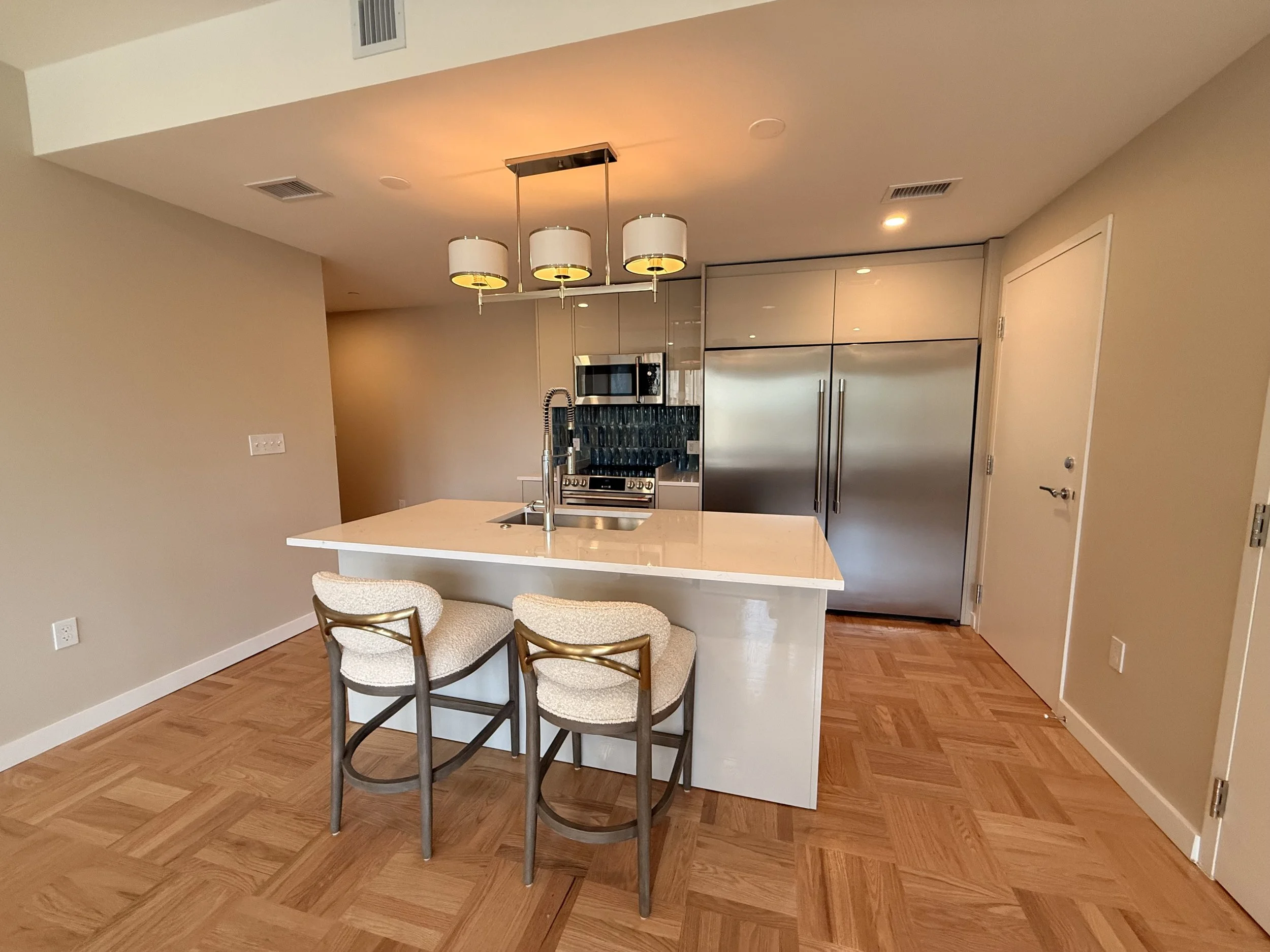 Modern kitchen with a white island, two beige chairs, stainless steel refrigerator, and beige walls.