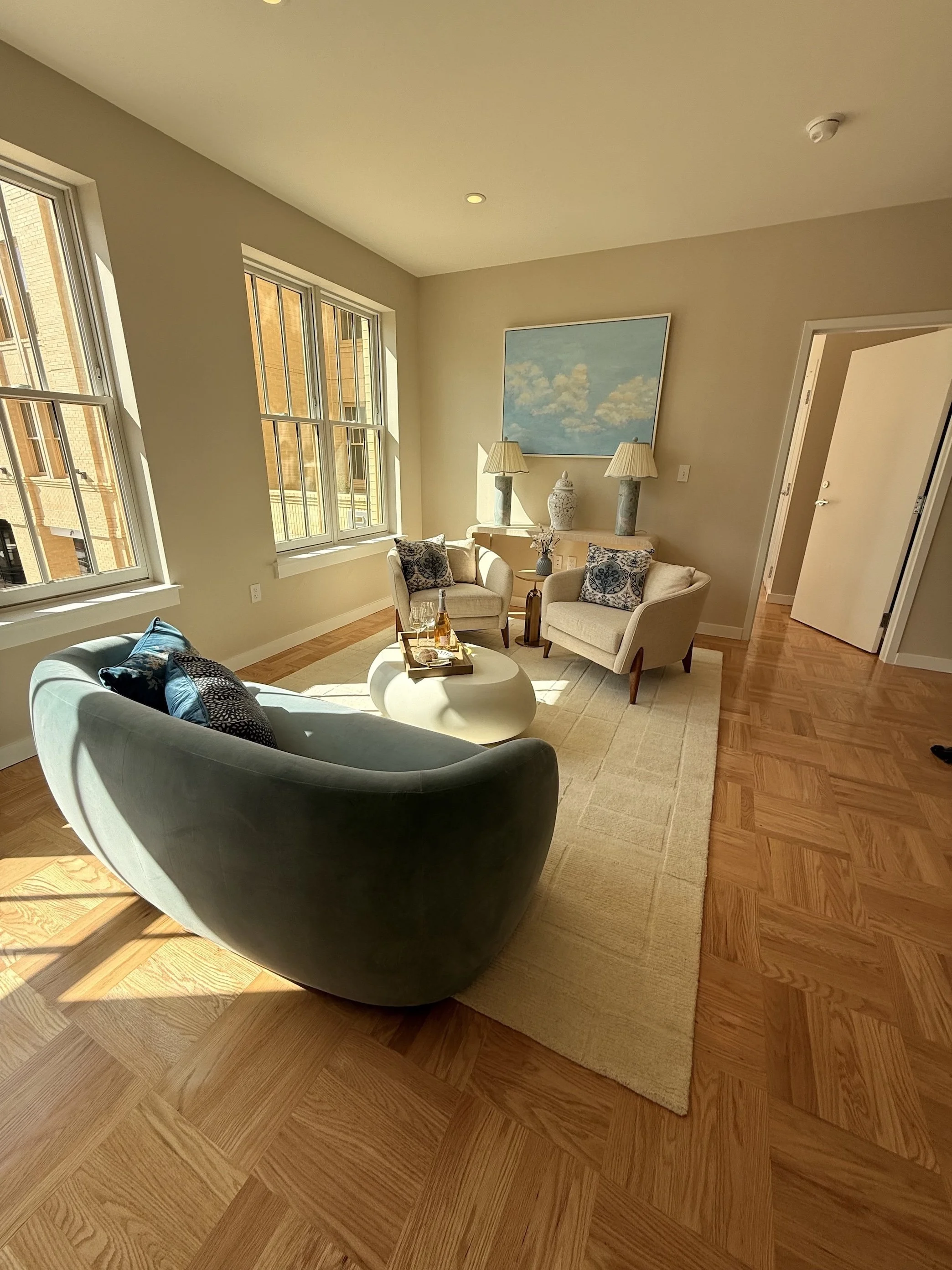 A bright living room with large windows, a curved sofa, two armchairs, a round white coffee table, and a painting with two table lamps on a console against the wall.