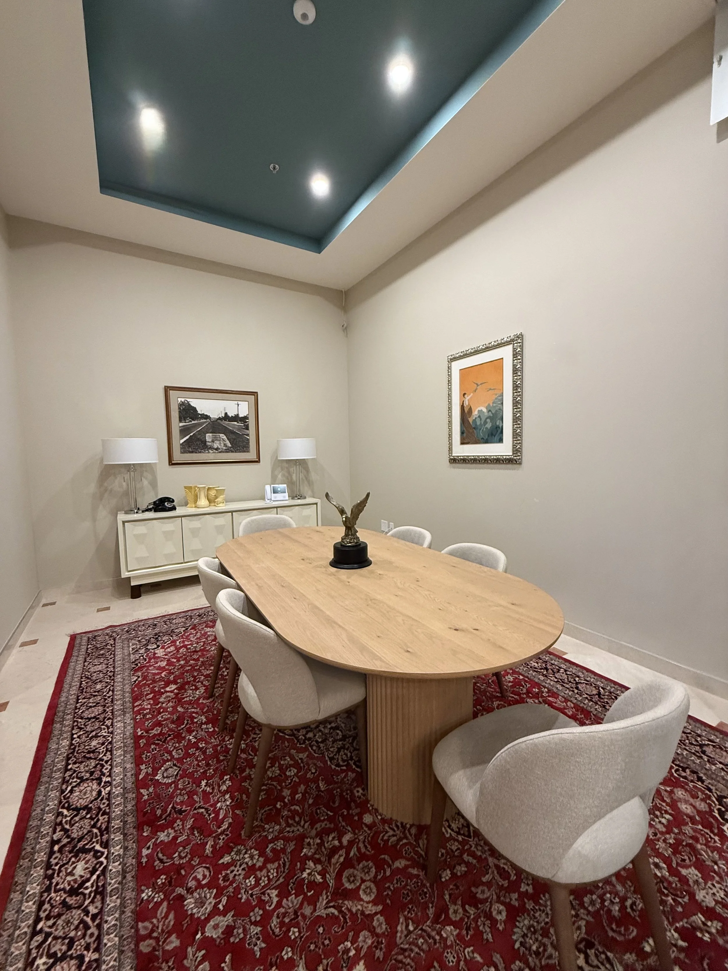 A modern dining room with a wooden oval table, six upholstered chairs, a red patterned rug, and a white sideboard. Two lamps and framed artwork are on the sideboard, and a sculpture of an eagle is on the table. The ceiling has a recessed blue section with three lights.