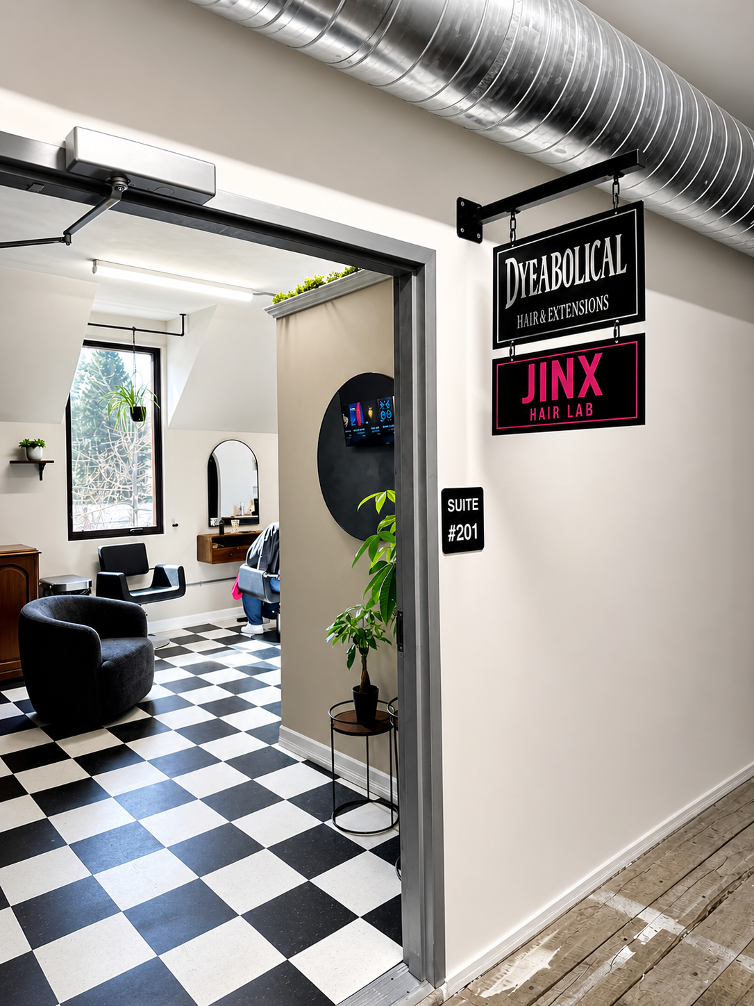 Interior of a hair salon with black and white checkered floor, black chairs, and a mirror. Sign hanging outside the door reads 'Dyeabolical Hair & Extensions' and 'JINX Hair Lab,' with a smaller sign indicating suite #201. There are plants, a window, and modern decor.