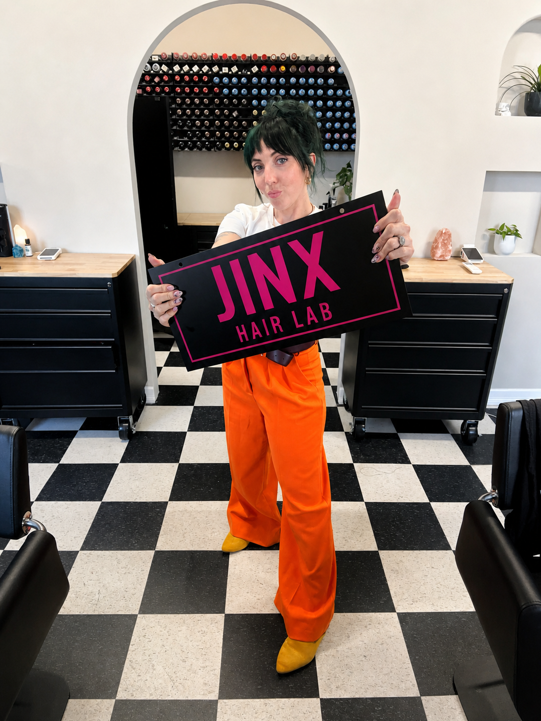 Woman with dark green hair holding a sign that reads 'JINX HAIR LAB' in a hair salon with black and white checkered floor and hair product wall in the background.