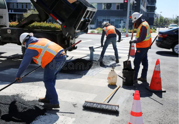 Construction workers in safety vests and helmets repairing a crosswalk on a city street, pouring asphalt and working near traffic cones.