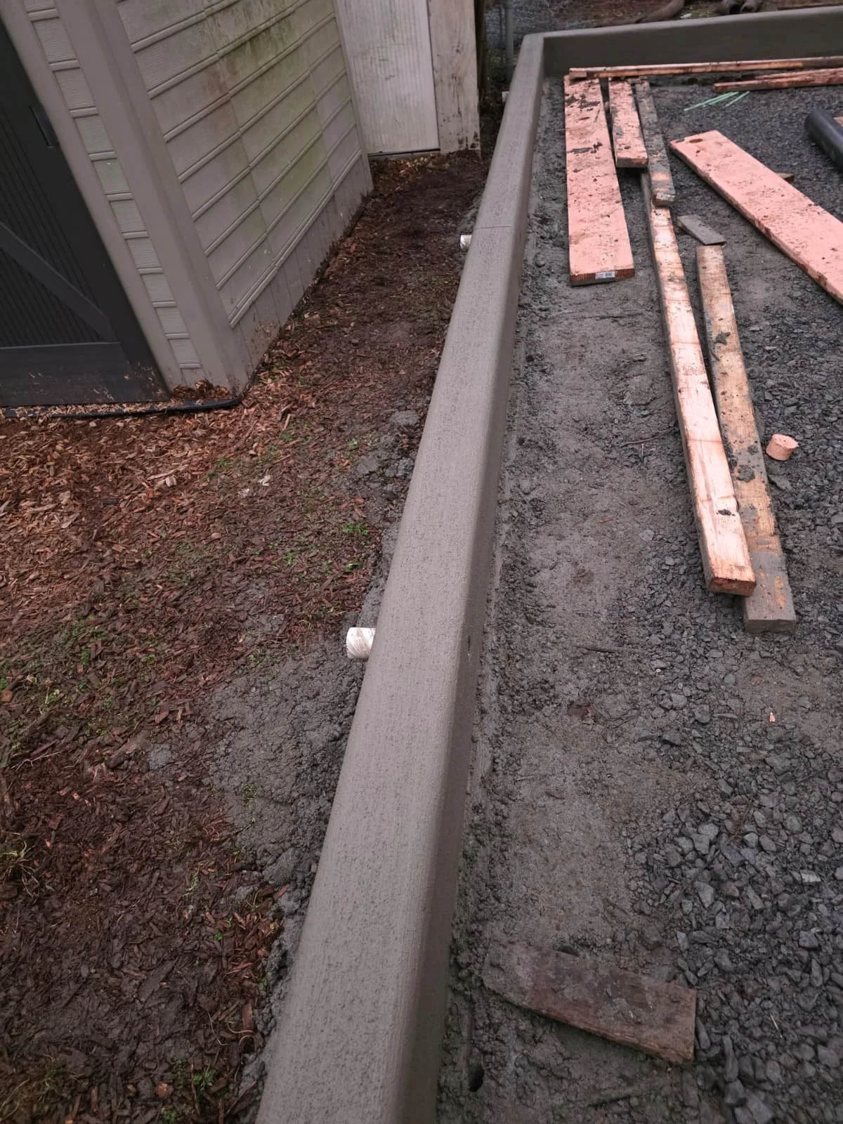 Construction site with a freshly poured concrete curb and wooden planks on gravel ground next to a house with gray siding.