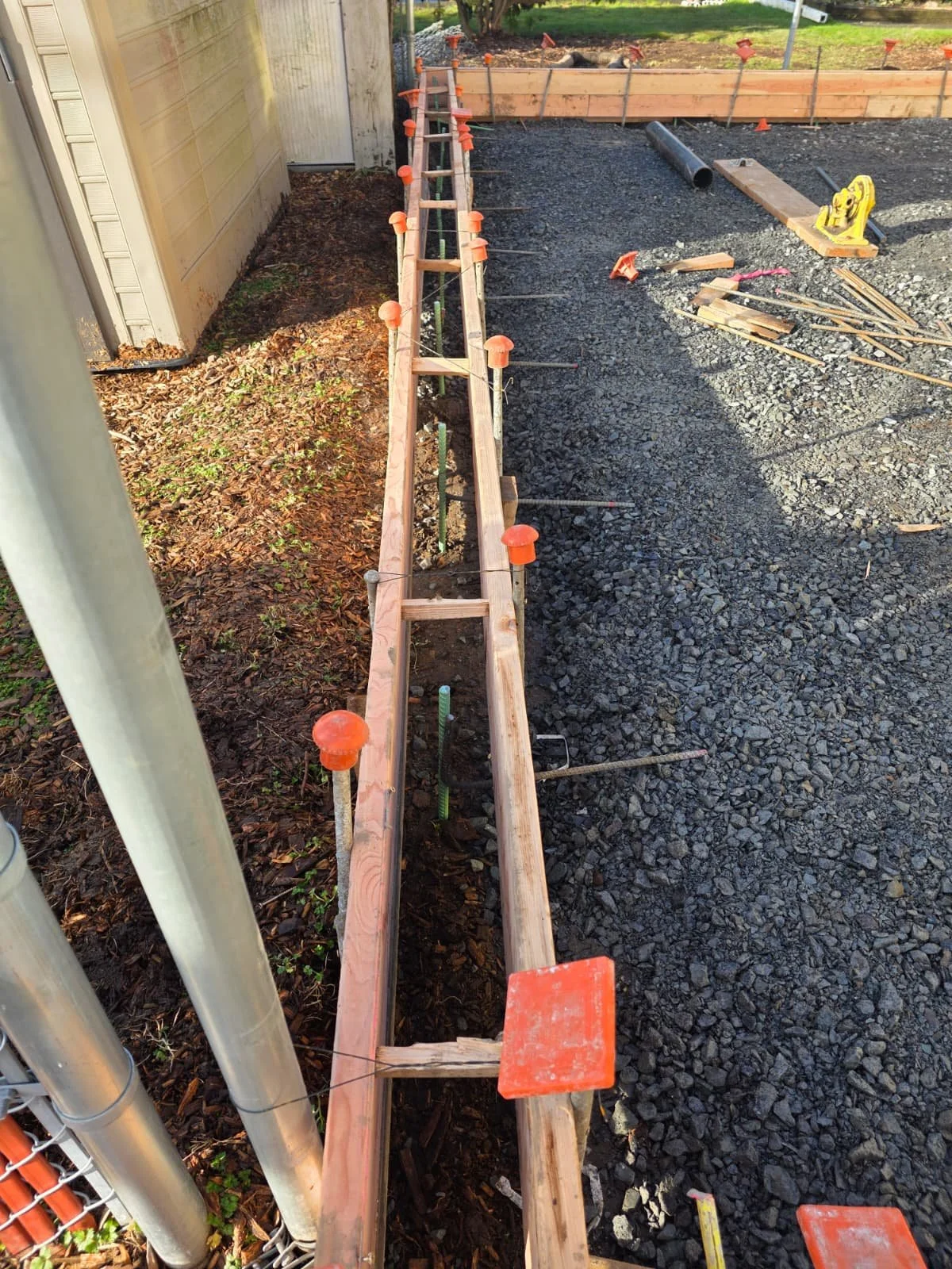 Construction site with wooden formwork and orange caps for concrete pouring, gravel ground, tools, pipe, and fencing in background.