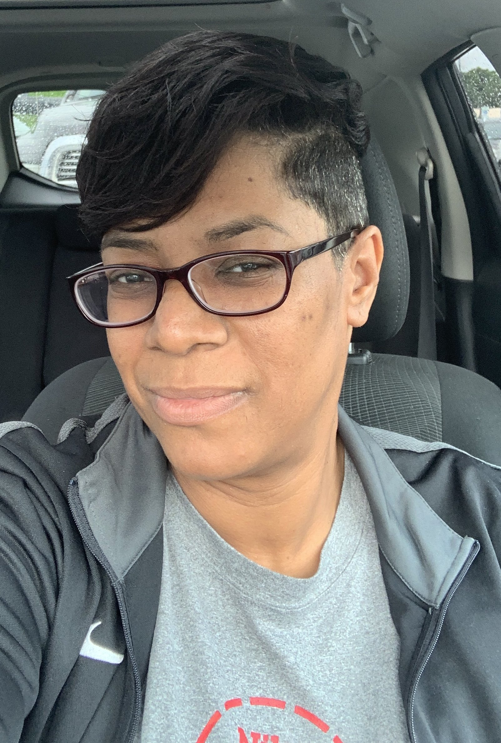 A person with short, styled dark hair, wearing glasses, a gray t-shirt, and a black jacket, sitting in a car, with trees and other vehicles visible through the windows.
