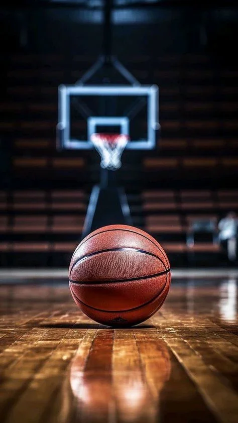 A basketball on a wooden court in a gym, with a basketball hoop and backboard in the background.
